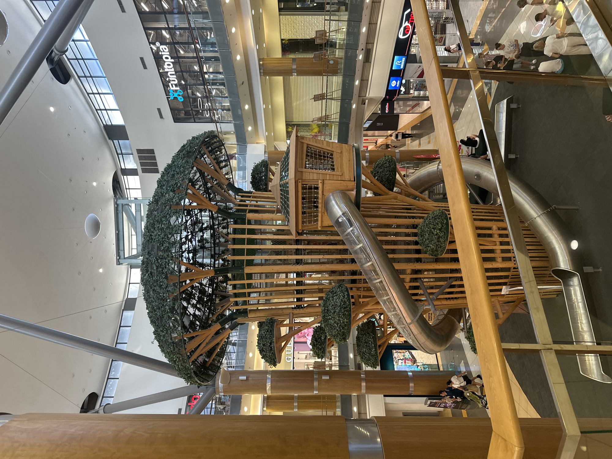 The image shows a large indoor playground structure resembling a tree, located in the central atrium of a multi-level shopping mall. The structure features wooden supports, artificial foliage, and a spiral metal slide. A small wooden cabin is integrated into the upper part of the tree structure. Various shops with illuminated signage are visible on different levels of the mall, including "Funtopia" and "Opticstore." People are scattered throughout the mall, some walking, some sitting, and a few are near the playground. The mall has a modern design with high ceilings, exposed beams, and glass railings. The overall lighting suggests an interior environment with natural light coming from large windows at the top and artificial lighting throughout.