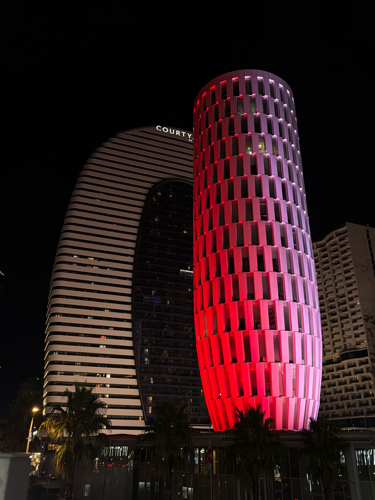A night view of the Batumi Marriott Hotel, located in Batumi, Georgia, reveals its distinctive architecture. The hotel is characterized by two connected buildings: one with a curved facade featuring horizontal white and dark stripes, and an adjacent cylindrical tower illuminated in vibrant shades of red and pink. The cylindrical tower's exterior is segmented by vertical elements that create a pattern of illuminated panels. The building to the left displays the "COURTY" logo in illuminated lettering. Palm trees are visible in the foreground, and streetlights and other buildings can be seen in the background, contributing to the urban night scene. The sky is dark, indicating nighttime.