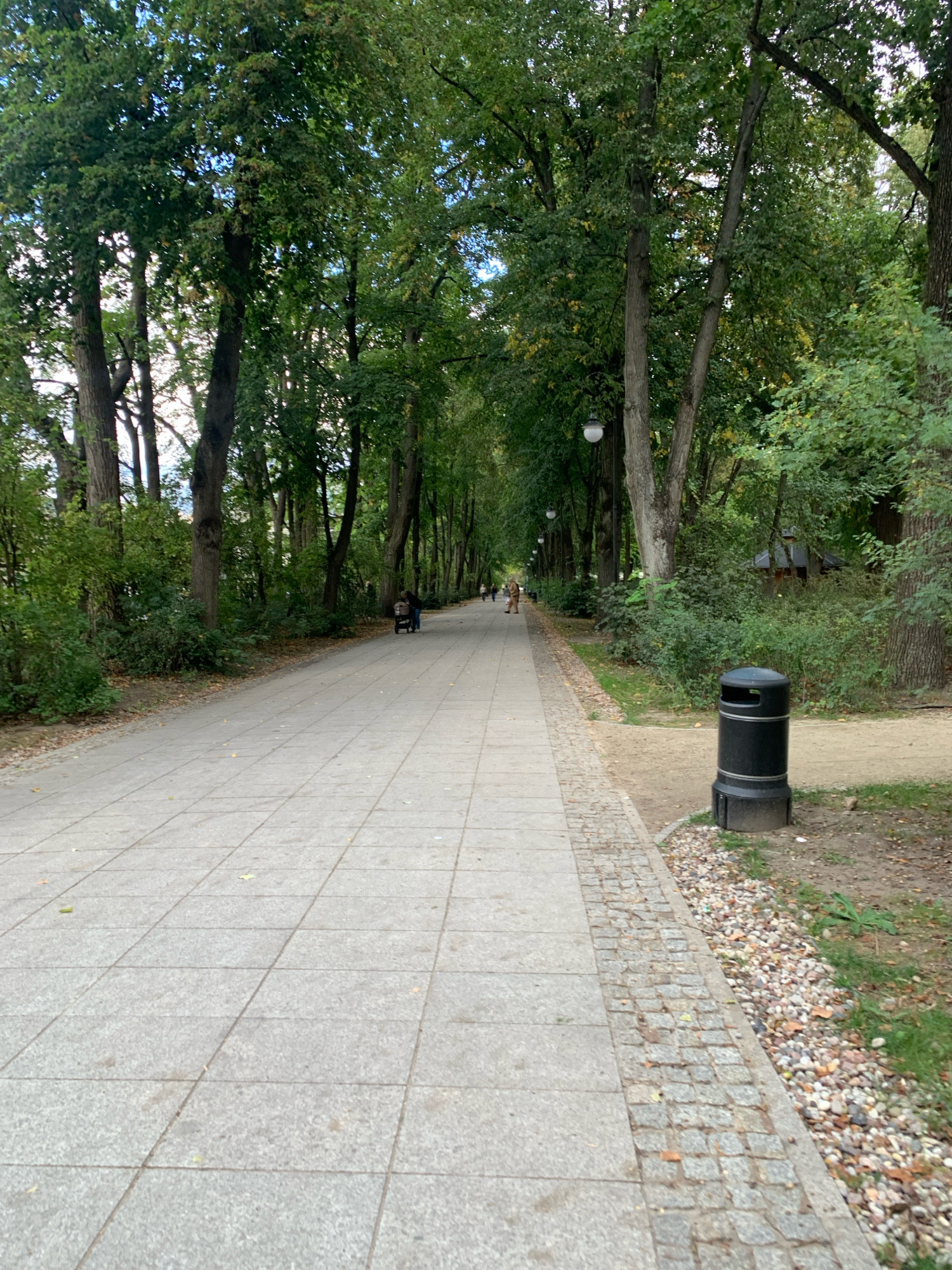 The image depicts a paved pathway in a park-like setting. The pathway, made of large, light-colored square tiles, runs straight into the distance, bordered on the left by lush green trees and foliage, and on the right by a strip of cobblestones, followed by a dirt path and grass. Several mature trees with dense green canopies line both sides of the pathway, creating a shaded avenue. A black trash can with silver trim stands near the right edge of the paved path. In the background, several individuals are walking along the pathway, and one person appears to be pushing a stroller. The overall atmosphere suggests a tranquil day in a public park. This scene is located in Planty Park in Białystok, Poland.