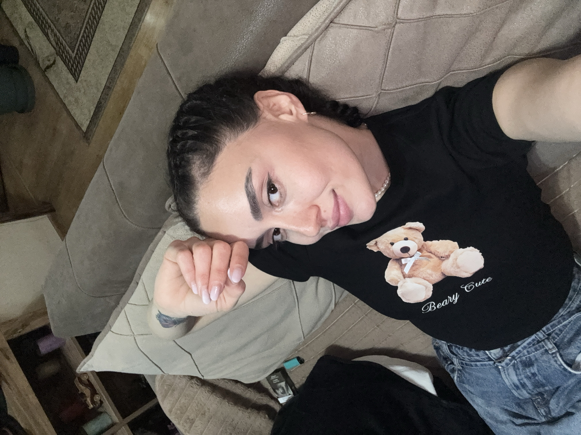 A woman lies on a couch. She wears a black T-shirt with a teddy bear and the words "Beary Cute" on it, and blue jeans. She has dark hair braided near her face and a pearl necklace. Her right hand rests on a pillow. The couch is brown and patterned, and a rug is visible in the background. There is no information available in the image to establish the location in Tbilisi, Georgia.