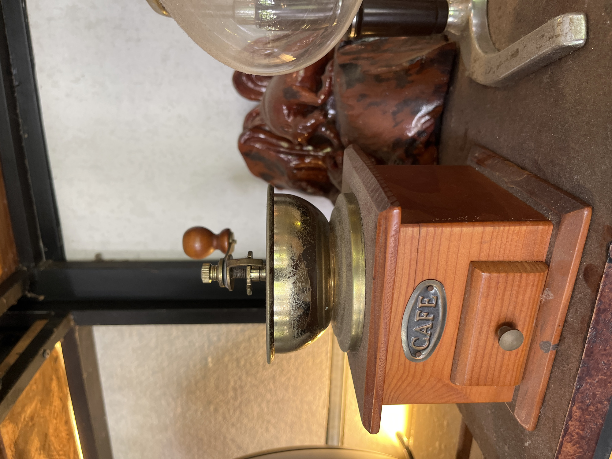 A high-angle, close-up shot shows a vintage coffee grinder with a wooden base and a brass grinding mechanism. The grinder has a drawer with a metal knob and a metal label that says "CAFE". The image also includes other decorative objects: a lightbulb, a carved wooden figure, and what appears to be part of a metal scale. The items sit on a surface with varied textures. The image lacks identifiable people and the exact location in Bac Nha Trang Ward, Vietnam, is not ascertainable.