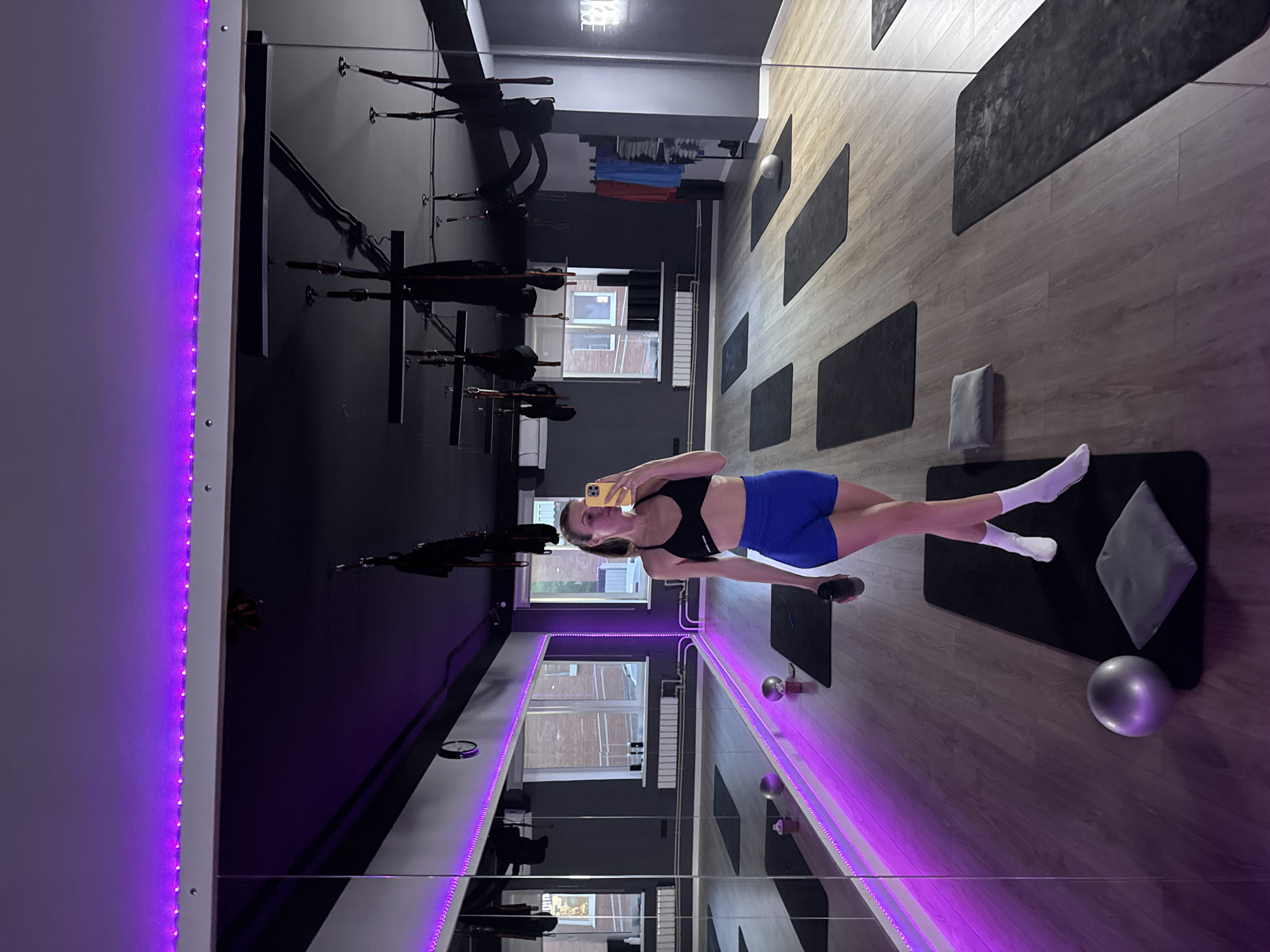 A woman in athletic wear poses for a selfie in front of a large mirror in a fitness studio. The studio has wooden floors, black walls, and purple LED lighting along the mirror's edge. Exercise mats are laid out on the floor. There are exercise tools suspended from the ceiling. Windows are visible in the background, showing an exterior view of brick buildings.