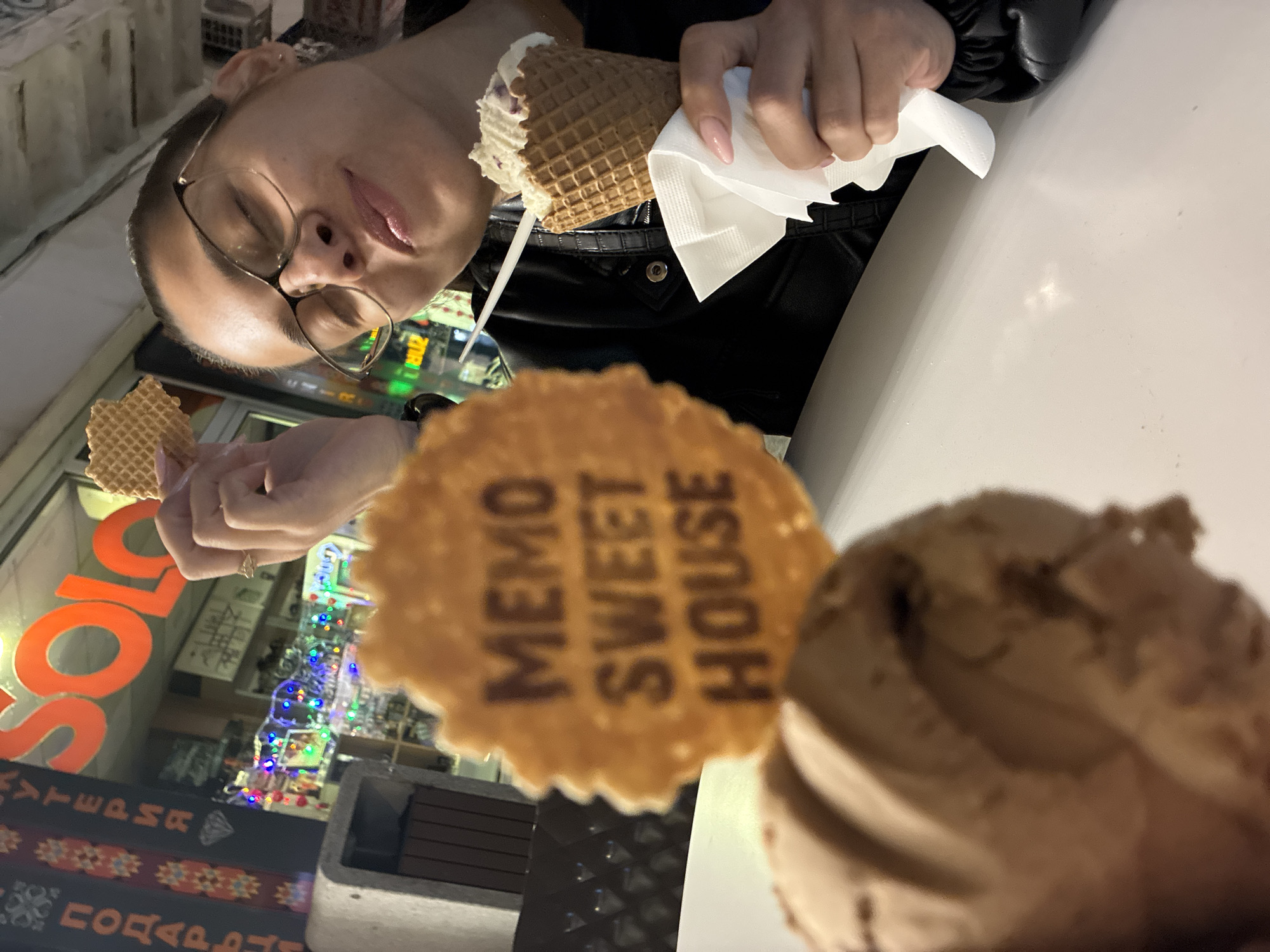 A person with glasses holds an ice cream cone wrapped in a white napkin in their right hand. In their left hand, they hold a small waffle. In the foreground, a scoop of ice cream is adorned with a "MEMO SWEET HOUSE" branded waffle. The environment appears to be a dessert establishment in Plovdiv, Bulgaria, as indicated by the storefront reflection in the table showing "SOLO" and part of the phrase "WATERMARK PODARAK." Christmas lights decorate the shop's interior. The person is wearing a black jacket.