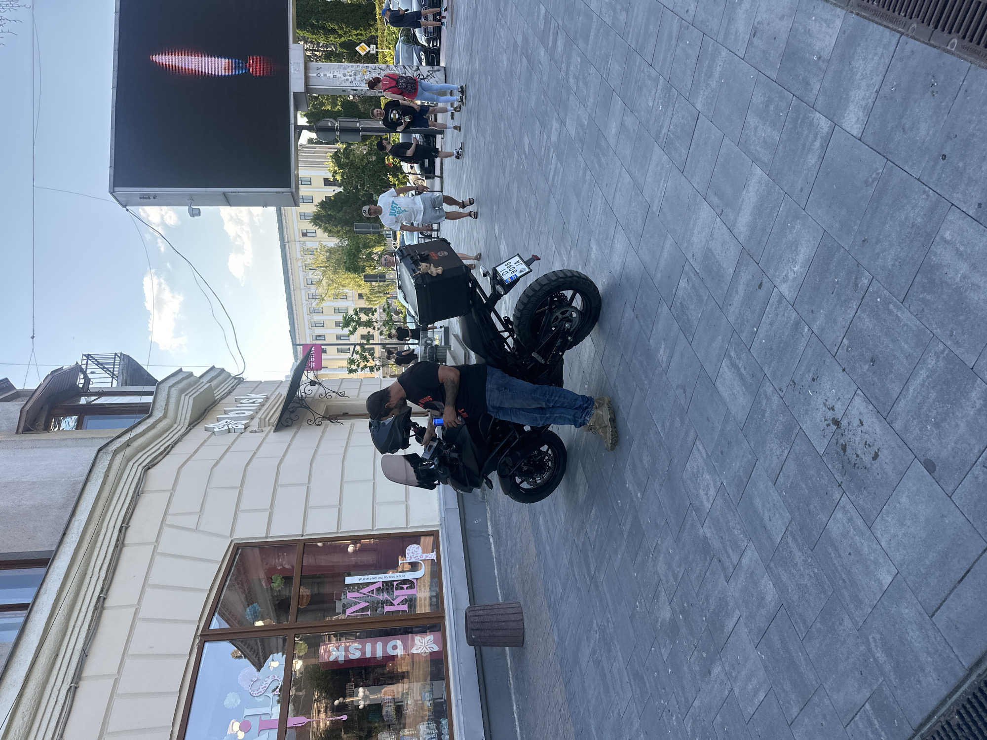 In Ternopil, Ukraine, on a sunny day, a man stands next to a black motorcycle on a wide sidewalk paved with grey rectangular tiles. The man, wearing a dark t-shirt, blue jeans, and beige shoes, holds a blue object in his hand. The motorcycle is equipped with a large black storage box on the back and a windshield. Behind the motorcycle, a building with a light-colored facade and large windows is visible, with "blisk" branding above an awning. In the background, other people are walking, some with backpacks, and cars are parked along the street. A large digital billboard displaying a red and white image of a candle with a flame is prominent in the background. The scene appears to be a public street or plaza in a city.