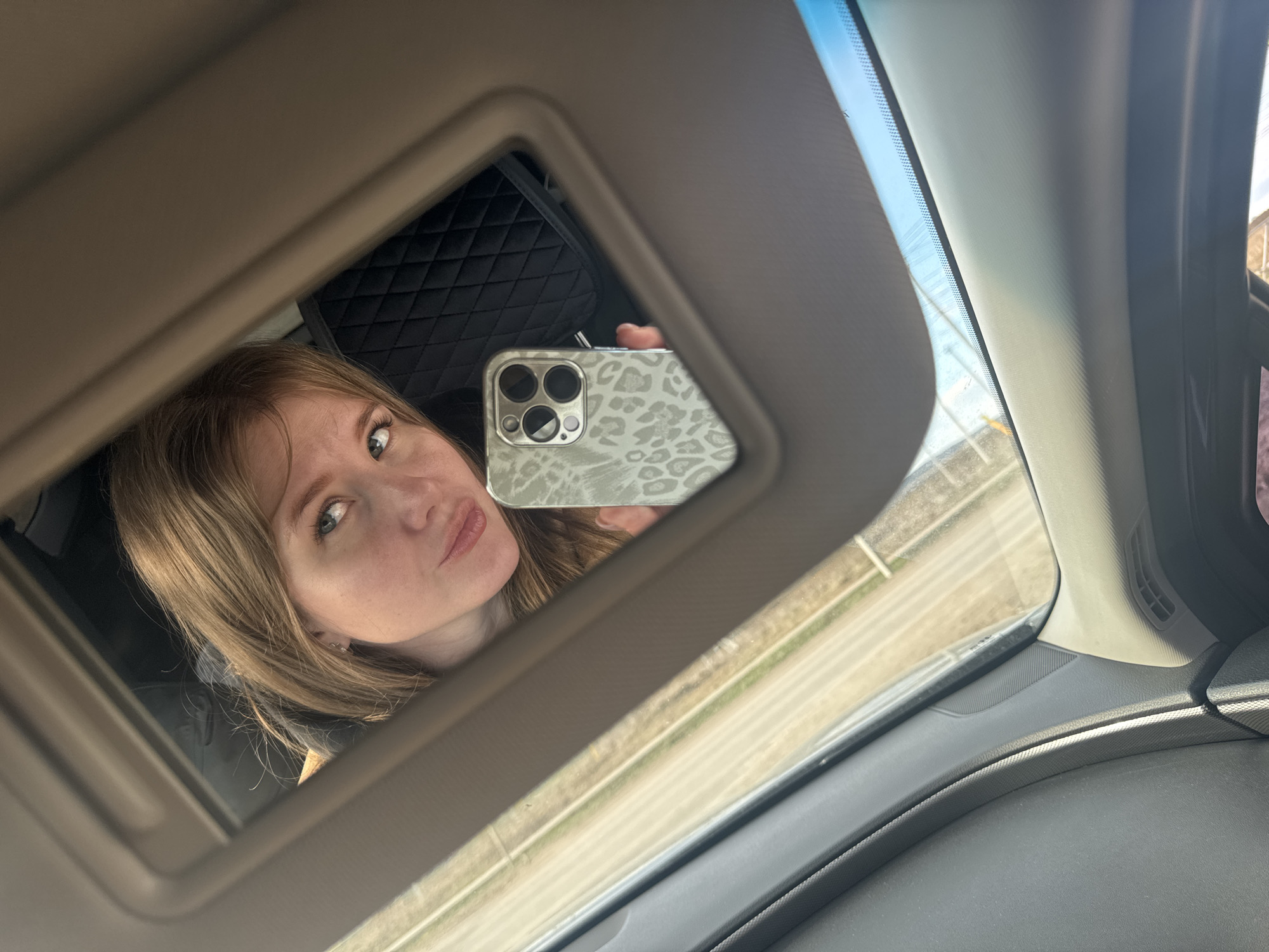 This image captures a selfie taken by a woman reflected in a car's open vanity mirror. The woman has long, light brown hair and is seen from the chest up, with her face prominently displayed in the mirror. She has light-colored eyes and her lips are slightly pursed, giving her a pouting or playfully serious expression as she looks directly into the phone's camera, which is reflecting in the mirror.
She is holding a smartphone, clearly visible within the mirror's reflection, which features a distinctive light-colored case with a speckled or leopard print pattern. The phone's multi-lens camera array is also visible. The immediate setting is the interior of a car, indicated by the vanity mirror (part of a sun visor), the windshield, a section of the dashboard, and a textured A-pillar. Behind the woman's reflection, a dark, quilted material, likely a car seat or headrest, can be seen.
Beyond the car's windshield, the outdoor environment is bright and sunny, suggesting it is daytime. A road or highway with a clear white line is visible, bordered by a light-toned, possibly grassy or sandy, shoulder. The sky appears clear. The overall scene suggests a casual moment during travel, possibly a road trip in the Горный, Russia region. No visible text is present in the image.