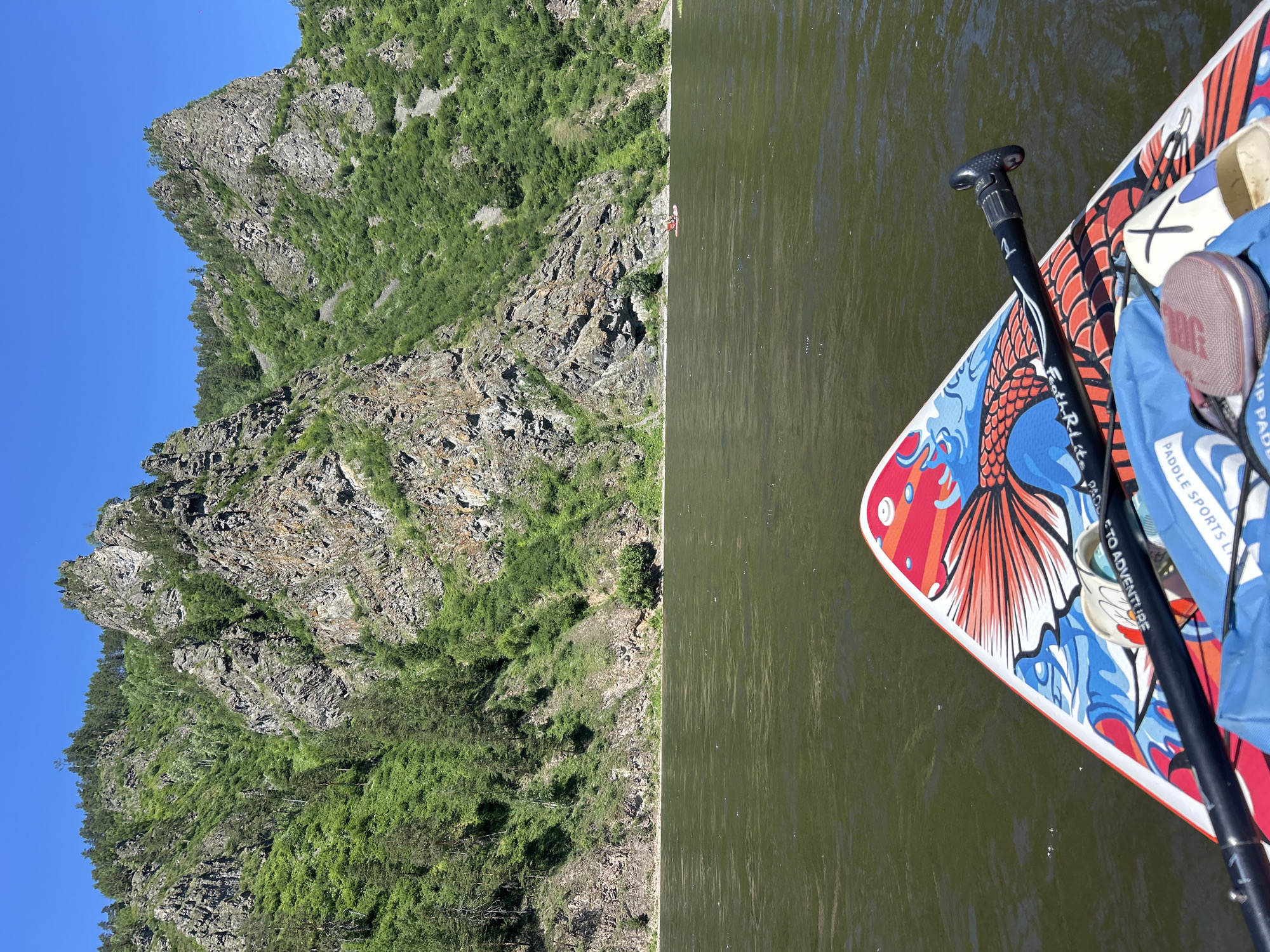 The image shows a paddleboard's nose and paddle on a river with a steep, rocky mountainside covered in green foliage in the background under a clear blue sky. The paddleboard is decorated with a colorful design featuring a fish and waves. A blue bag and a round, mesh-covered object are also visible on the paddleboard. In the distance, on the river, another person is visible on a small red raft. The environment suggests a natural, outdoor setting, possibly a recreational area. The location is identified as Ust-Mana, Russia.