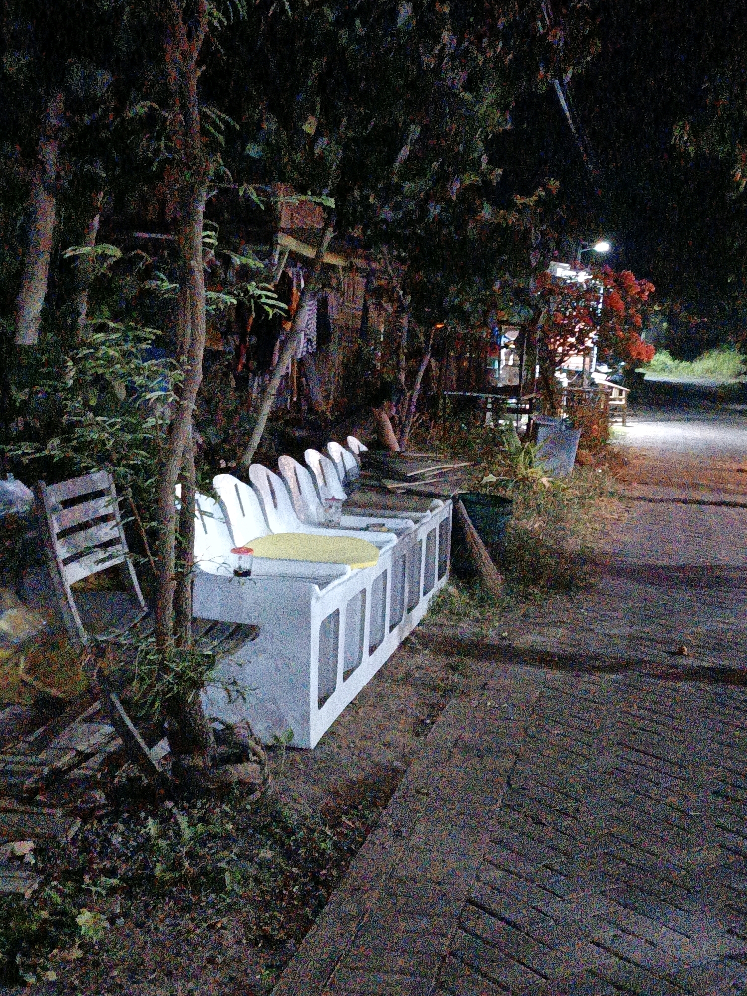 The image shows a row of white concrete benches with yellow seats in what appears to be an outdoor seating area. The benches are arranged in a line, with one end of the row cut off from view. A wooden chair is placed to the left of the benches, slightly angled towards the foreground. In the background, there are trees and foliage, with some laundry hanging from a line. A street light is visible in the upper right corner, illuminating a pathway or road made of dark paving stones. The scene is dimly lit, suggesting it is evening or night. The environment appears somewhat overgrown with vegetation. There are no people visible in the image. The location is identified as Cikal, Indonesia.