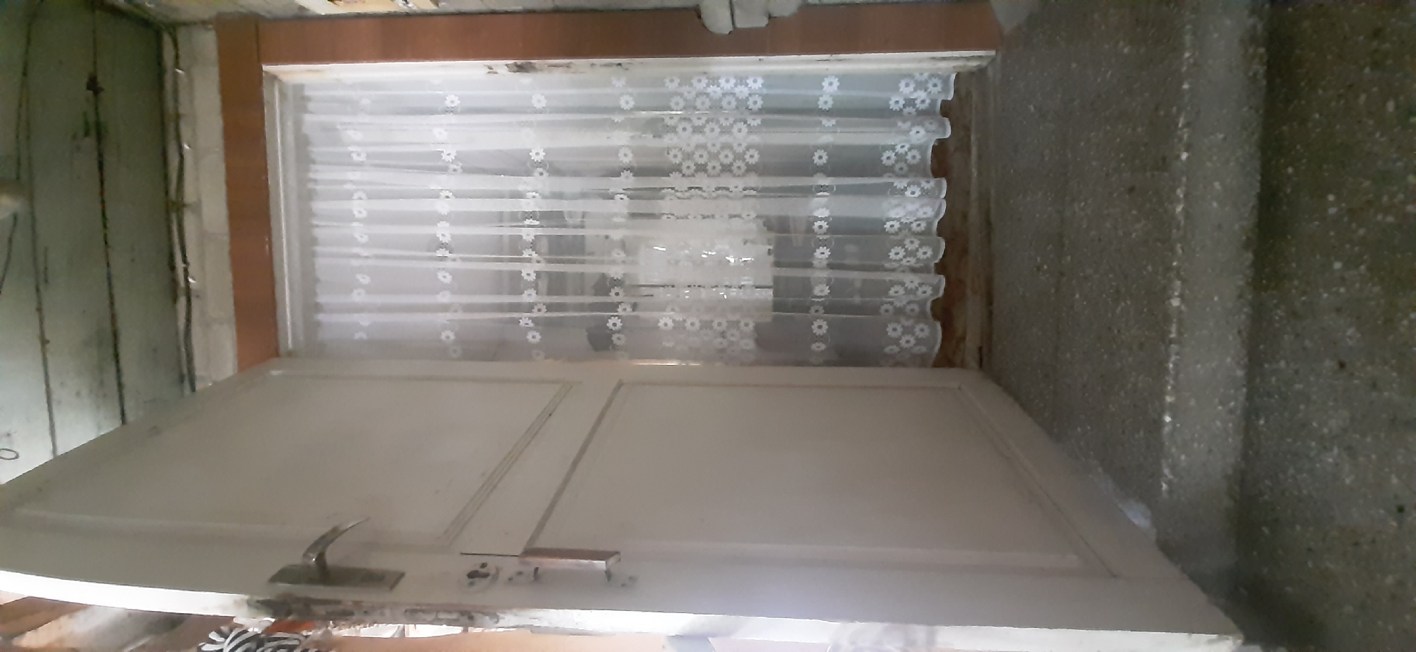 The image shows the interior of what appears to be a doorway or hallway in a residential setting. The main subject is a white, two-panel door with a silver handle and lock mechanism. Above the door, a sheer white curtain with a delicate floral pattern is visible, suggesting a window or glass panel. The curtain is pleated, creating a textured effect.
To the left of the door, there's a wall with a distressed, possibly wooden, finish, exhibiting vertical lines and some wires or conduits running along it. To the right of the door, a textured concrete wall is present, with a speckled pattern of darker and lighter aggregates. The floor in the foreground and right side of the image is not fully visible but appears to be made of concrete or a similar rough material.
The scene appears to be indoors, likely in a home or a similar structure. There is no indication of specific time of day or weather conditions, and no people are visible. The lighting is somewhat dim, consistent with an indoor environment, possibly illuminated by artificial light or indirect natural light. The overall impression is of a utilitarian or somewhat aged interior space. No text is visible in the image.