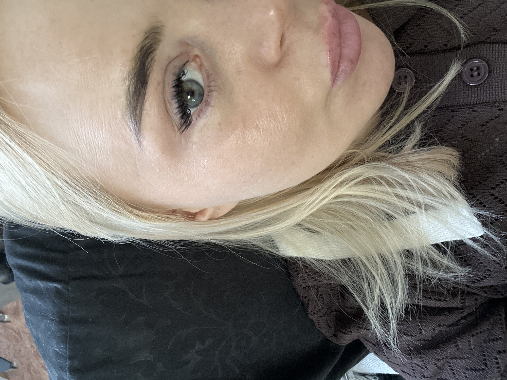 A close-up, high-angle shot captures the right side of a person's face, oriented horizontally. The individual has light blonde hair, which cascades down around their head. Their skin is fair, and a prominent dark eyebrow frames a light blue-green eye. The eye features long, dark eyelashes, possibly enhanced with mascara or extensions. Fine lines are visible on the skin around the eye and forehead.
The person's nose is partially visible, and their full lips have a glossy, pinkish tint. They appear to be lying down, possibly on their side. A dark brown garment, which seems to be made of a textured, possibly knitted or lacy material, is visible around their chin and shoulder area. Two dark purple buttons are noticeable on this garment. Beneath their head, there is a dark fabric with a subtle, dark patterned design, possibly a pillow or another piece of clothing. No text is visible in the image.
