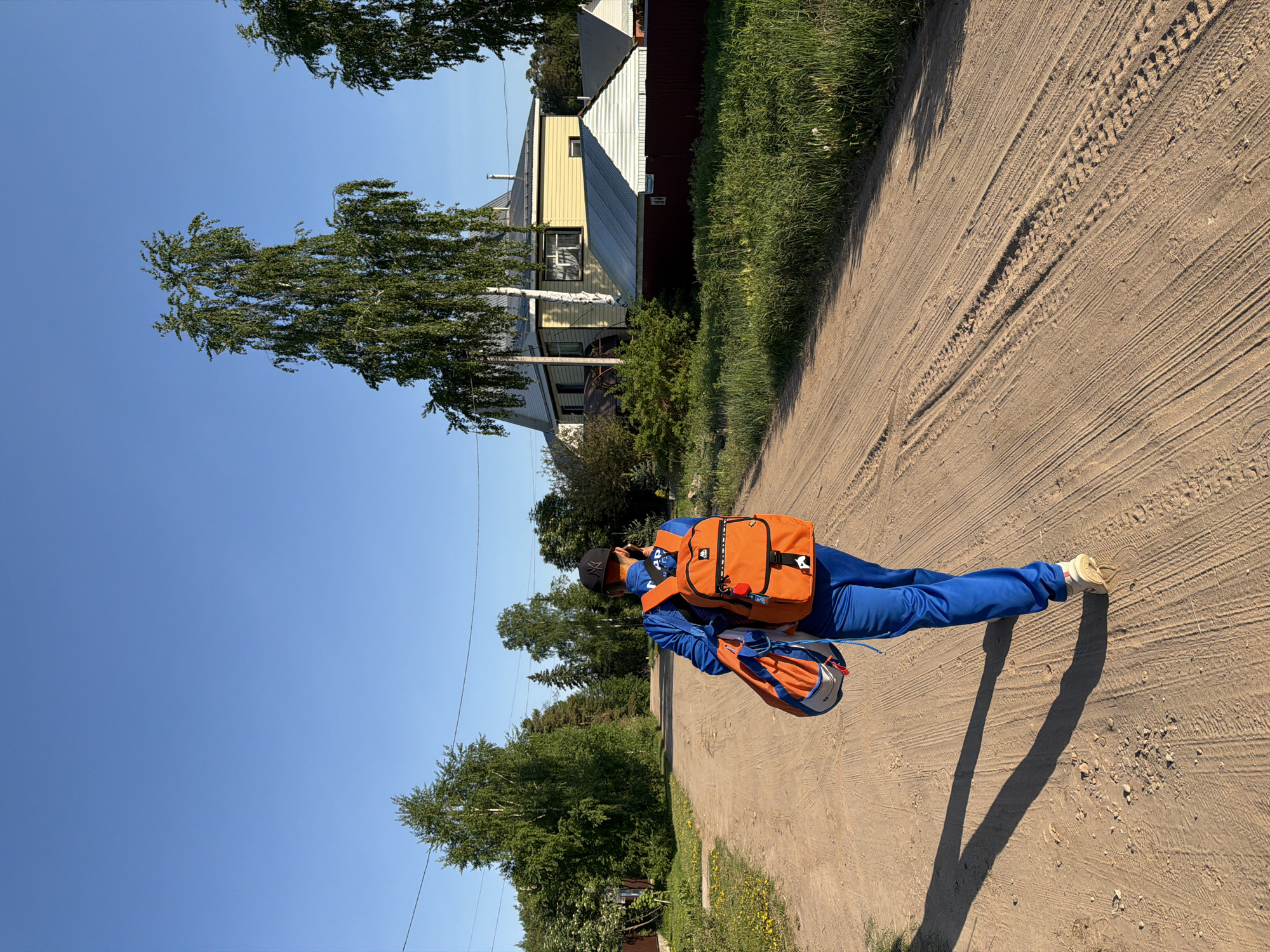 The image shows a person wearing a blue tracksuit, a black baseball cap, an orange backpack, and light-colored shoes standing on a dirt road. They are oriented at an angle to the viewer. The dirt road has tire tracks and leads into the distance. To the left, there are trees and a clear blue sky. To the right, there is a building with a yellow and white exterior and a dark-red roof. The location is determined to be in the city of Chaikovsky, Russia, based on analysis of the surrounding environment and architectural style, which is typical of residential areas in that region.