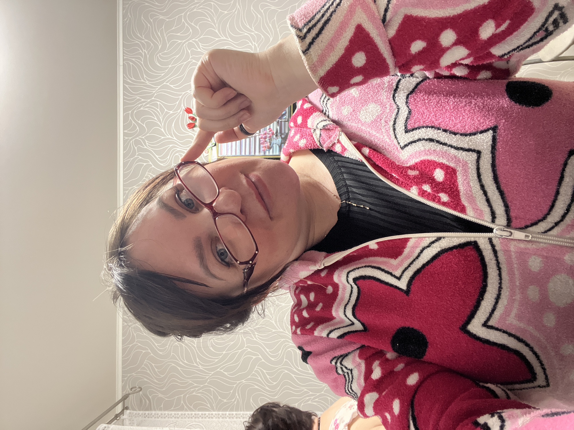 A person wearing eyeglasses and a pink patterned robe points to their forehead in an indoor setting. The patterned wallpaper and visible portion of a towel rack suggest a bathroom or bedroom. In the lower portion of the image, another person’s head is partially visible. Location in Perm, Russia cannot be determined from the image.