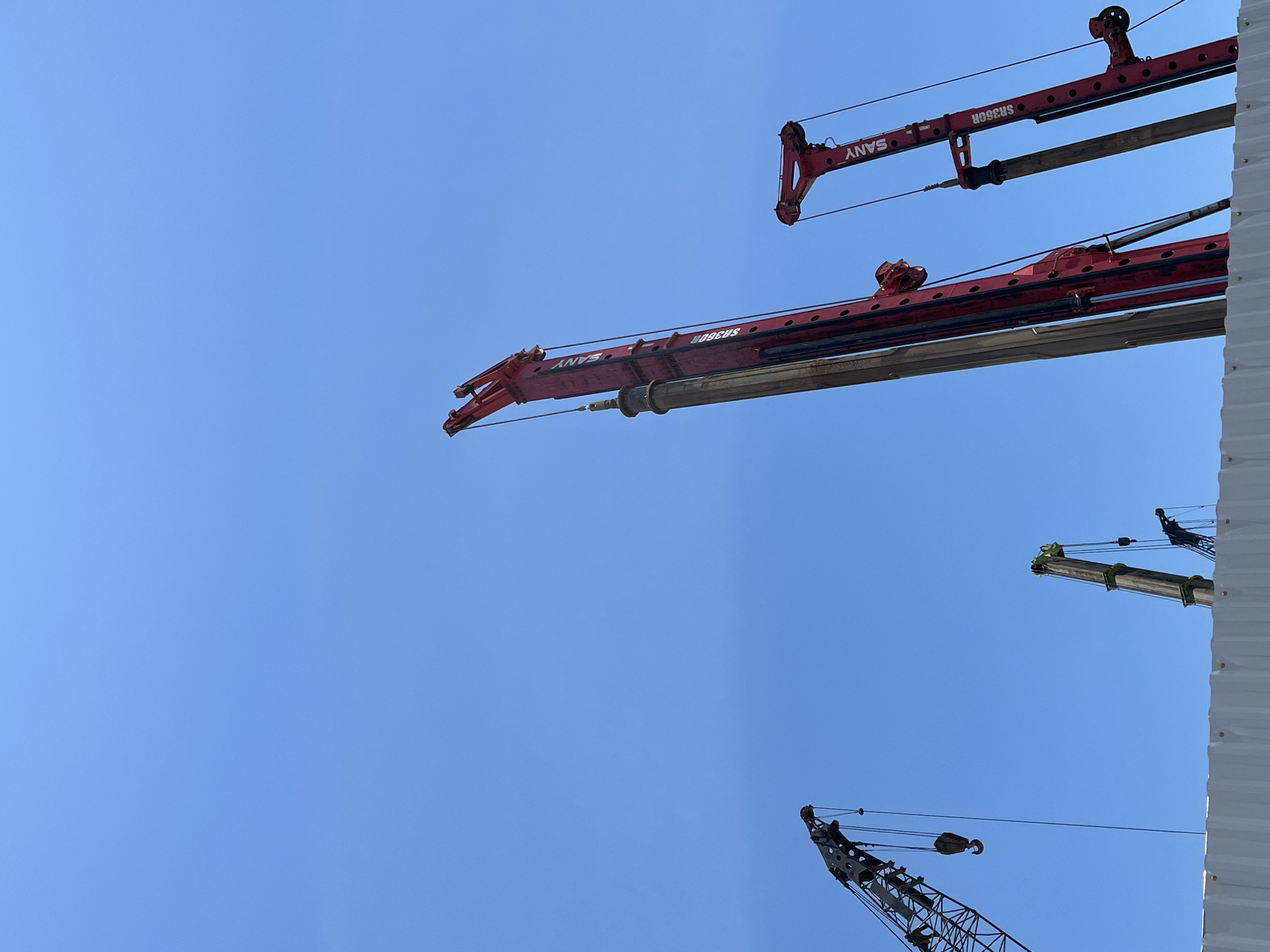 The image shows a construction site in Đà Nẵng, Vietnam. Visible are the booms of several construction cranes against a clear blue sky. Two red cranes, labeled "SANY SR360R", are prominent in the upper part of the frame. Other crane booms appear toward the bottom and right edge. A white, corrugated metal structure is visible on the right side. No people are visible in the frame.