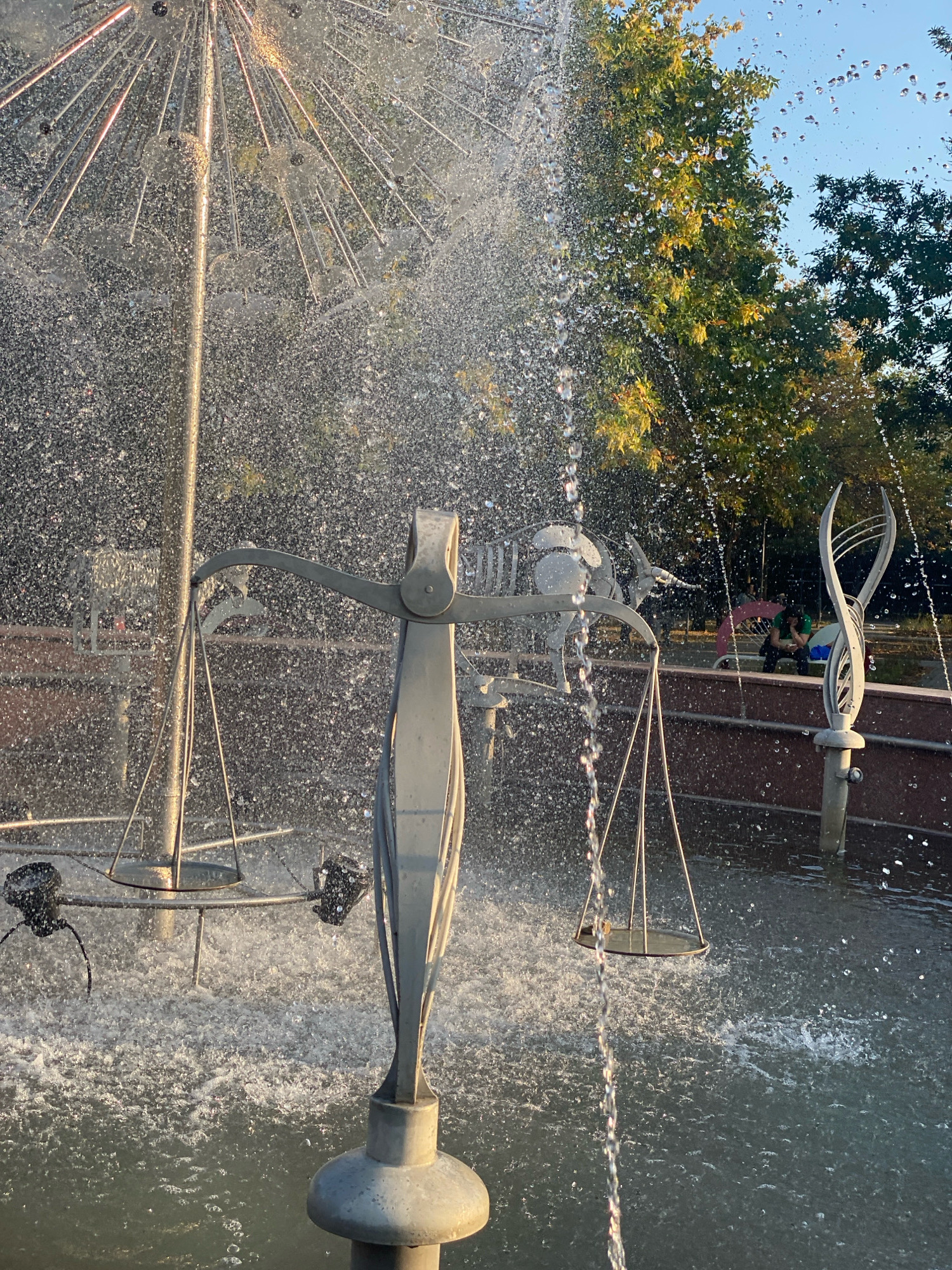 The image captures a close-up view of a fountain sculpture, likely located in Chișinău, Moldova, given the context. The sculpture features a stylized set of scales, a common symbol of justice, with metallic pans suspended from its arms. Water sprays from various points of the fountain, creating a dynamic and shimmering effect as droplets catch the sunlight. In the background, lush green trees with hints of yellow foliage suggest an autumn season. The sky above is a clear blue. Further in the background, blurred figures of people can be seen, some seated on a bench, one holding a red umbrella, indicating a public park or recreational area. The overall impression is one of a tranquil outdoor scene, where art and nature converge, with the energetic play of water adding a lively element.