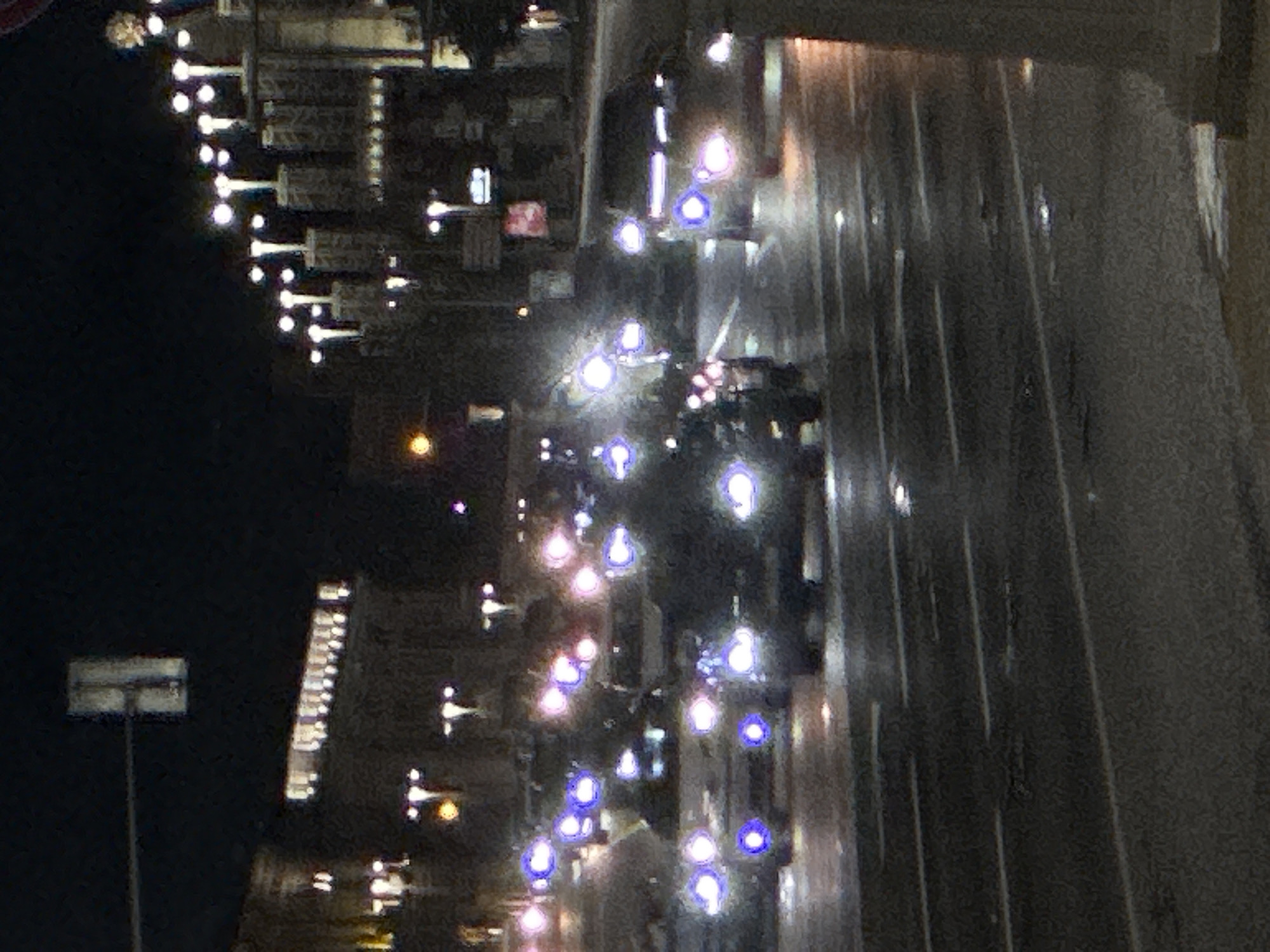 The image captures a nighttime scene on a city street, likely in a downtown area, with wet asphalt reflecting the lights of vehicles and buildings. Several cars are visible on the road, their headlights and taillights illuminated, creating streaks of light due to the motion blur and low light conditions. The foreground shows a wet street with lane markings. In the background, buildings with illuminated windows line the street. Traffic lights and streetlights are also present, adding to the urban atmosphere. The overall impression is one of a busy city environment at night, with vehicles navigating the wet road.