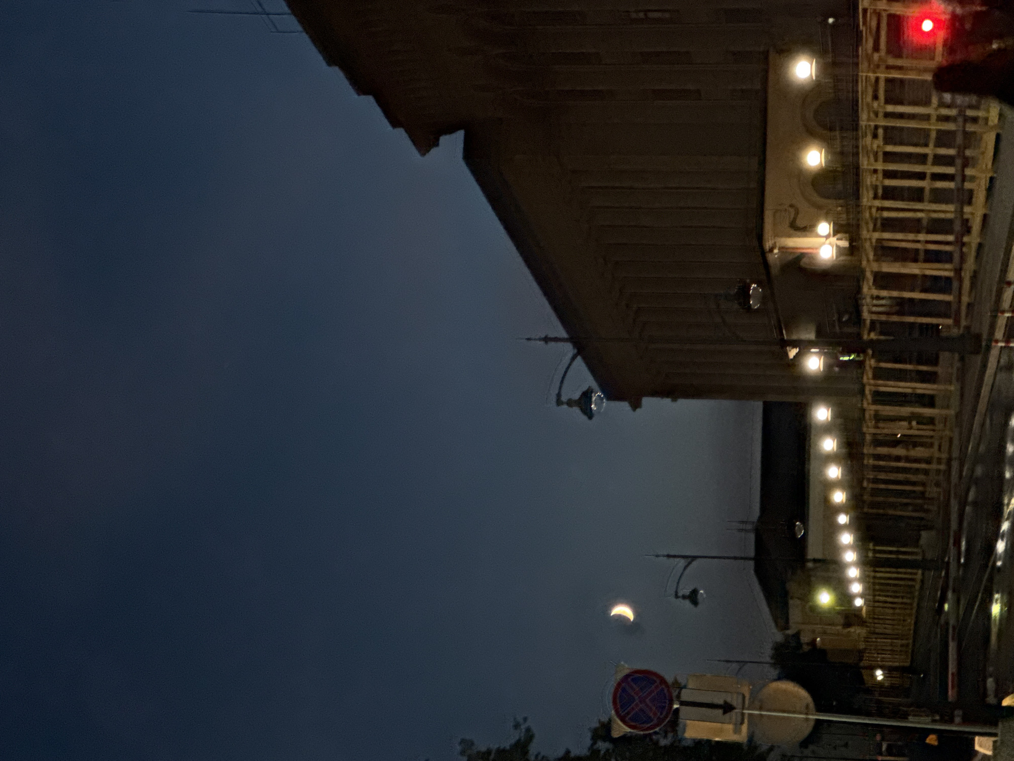 The image captures a nighttime scene on a street in Nizhny Novgorod, Russia, possibly near a historical building. The sky is a deep, dark blue, with a crescent moon visible. On the left side of the frame, a traffic sign with a red cross inside a circle and a white circle below it is mounted on a pole. A street light with a visible bulb stands nearby. To the right, a large, imposing building with a facade featuring vertical columns and arched windows is illuminated by streetlights. Along the street, a row of evenly spaced, glowing streetlights illuminate a wooden fence-like structure. The wet asphalt of the street reflects the lights. In the far right, a red traffic signal light is visible, and the silhouette of a person can be seen.