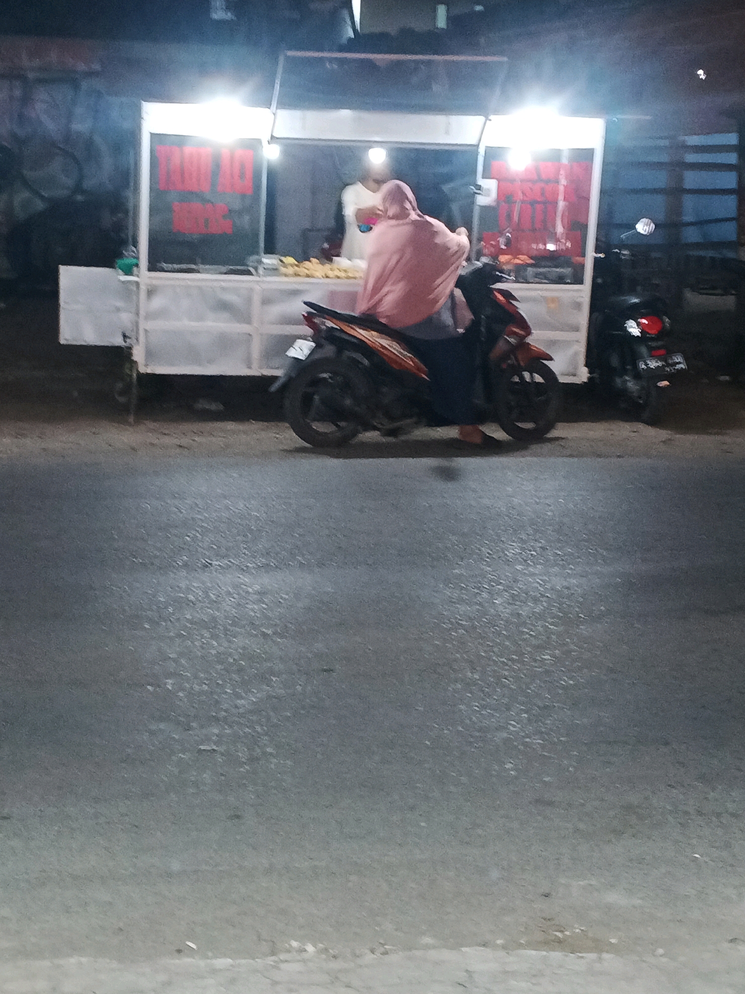 In Taman, Indonesia, a food stall is operating at night. The stall, which appears to be mobile with a white wheeled base, has bright overhead lighting illuminating its contents. On the left side of the stall, a sign with red text is visible, but illegible due to the image quality. The stall displays various food items, seemingly fried or grilled, in trays. Two individuals are present: one person, wearing a pink headscarf and dark clothing, is seated on a red and black motorcycle parked in front of the stall, facing away from the camera. This person is partially obscured by the motorcycle. Another person, wearing a white shirt, stands behind the counter, interacting with or serving the seated individual. To the right of the main stall, another motorcycle is parked. The background consists of a dark, somewhat textured wall, possibly graffiti-covered. The foreground shows a paved road surface, indicative of a street or parking area. The overall scene depicts a typical roadside food vending operation at night.