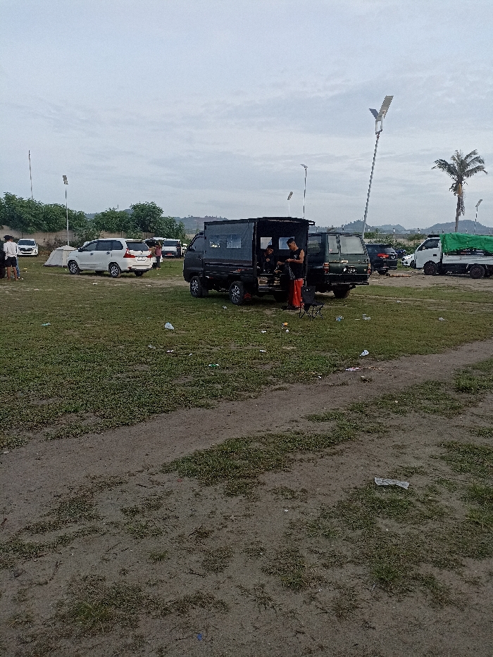 This image depicts an outdoor gathering of people and vehicles in an open, grassy field in Pujut, Indonesia, under an overcast sky. The lighting is soft and diffused, suggesting a cloudy day or the transitional hours of dawn or dusk.
In the foreground, a black pickup truck with an enclosed cargo bed is parked on the grass. Two men are visible near its rear; one, wearing a black t-shirt and orange pants, stands by the open cargo area, seemingly interacting with its contents, while another person is partially seen inside the truck. A small folding chair is placed beside the truck. Another similar black truck is parked behind it.
Further into the scene, several other vehicles are present, including a white Toyota Innova and other white cars, with various people standing around them. A white tent is pitched near these vehicles, suggesting a camping or outdoor leisure activity. In the distant background, a white truck with a green tarp-covered bed is also visible.
The ground is a mix of green grass and bare dirt paths, with some scattered litter indicating a casual, perhaps well-used, public space. Tall poles equipped with modern solar streetlights are intermittently placed across the area. A tall palm tree stands on the right, and lush, vegetated hills or mountains form the distant backdrop. The scene conveys a relaxed and informal atmosphere, consistent with a social outing or event in a semi-developed outdoor setting. No legible text is visible in the image.