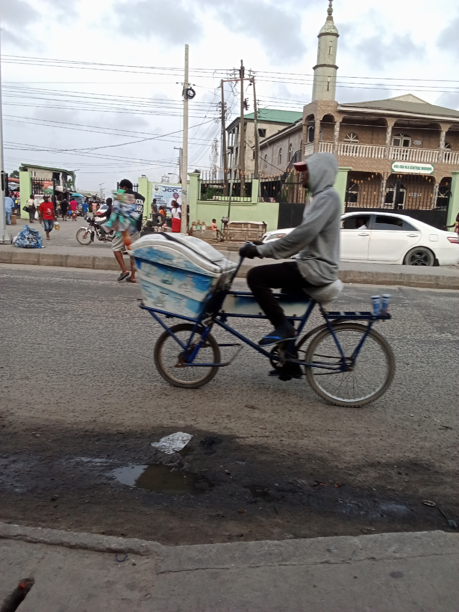 This image depicts a lively street scene in Ado, Nigeria, under an overcast sky. The primary subject is a person, dressed in a gray hooded sweatshirt, black pants, and flip-flops, riding a custom-built blue bicycle. This bicycle is modified with a large, light-blue and white cooler or container attached to the front, suggesting it might be used for selling refreshments or goods. Two small blue cans are visible on a rear rack.
The foreground shows the asphalt road, which has visible potholes and puddles, indicating recent rain or poor drainage. A concrete curb runs along the right side of the road.
In the midground, the scene is bustling with activity. Numerous pedestrians are visible, some walking towards the background, others closer to the viewer. A person in a red t-shirt stands near a large blue bag of refuse on the sidewalk. A motorcycle is also present, moving or parked among the people. Towards the right, a white sedan is parked beside the road. Behind it stands a prominent multi-story mosque, distinguished by its tall minaret and tan-colored facade with arched windows and balconies. A green sign on the mosque clearly displays the text "OKE-IRA MLA CENTRAL MOSQUE." The mosque compound is enclosed by a green fence or wall.
Further in the background, more buildings and dense overhead power lines crisscross the sky. Partially visible signage includes "JCC" and another sign with text that appears to be "UCKIN GARD." The cloudy sky provides soft, ambient lighting, suggesting either an early morning, late afternoon, or simply a heavily overcast day. The overall impression is a typical, busy urban environment in West Africa, characterized by everyday commerce and public movement.