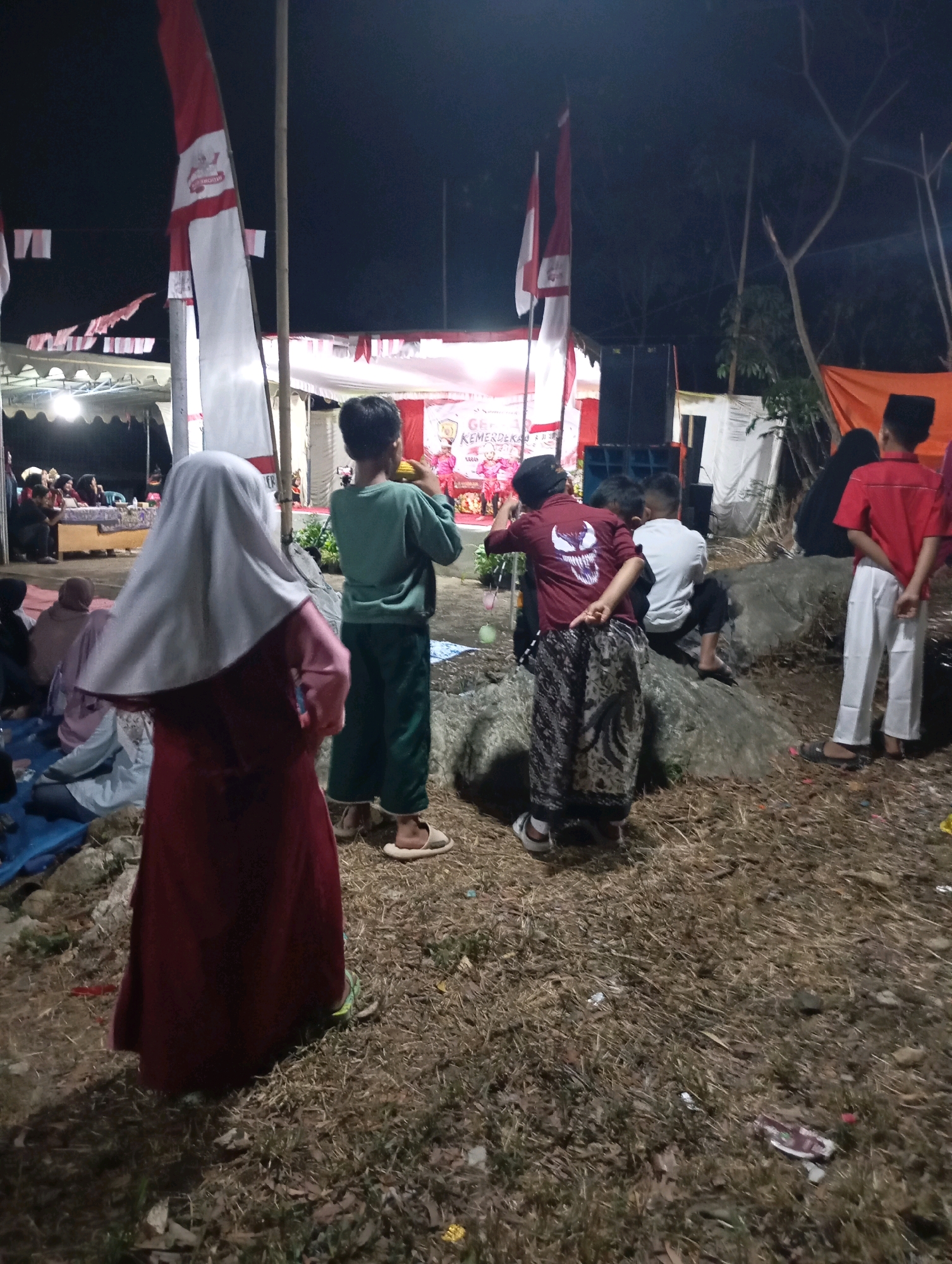 This image depicts an outdoor evening gathering in Wonogondo, Indonesia. The scene is lit by artificial lights, suggesting it's a nighttime event. In the foreground, a person wearing a maroon hijab and dress is partially visible from the back. To their right, a young boy in a green shirt and pants is holding a yellow object, possibly food. Further to the right, a person in a red shirt with a "Venom" logo and patterned pants stands with their back to the camera. Another child in a white shirt is seated on a rock. In the background, a stage area is set up with white draping and banners, some of which appear to be Indonesian flags. Speakers and tents are also visible, indicating a community event or celebration. The ground is covered with dry grass and some scattered debris. The overall atmosphere appears casual and communal.