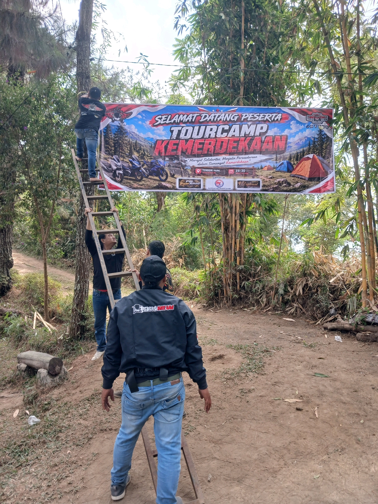 The image depicts an outdoor scene in Berjo, Indonesia, likely during a "Tourcamp Kemerdekaan" event. In the foreground, three individuals are present. One man in a black jacket with white lettering on the back is standing with his back to the camera, facing away from a ladder. His blue jeans and grey sneakers are visible. To his left, another man in a black shirt and blue jeans is holding onto a wooden ladder, looking up at someone above. His action suggests he is either assisting or observing the person on the ladder. A third person, partially visible, is also in the vicinity.
The central focus of the image is a large banner stretched between trees and bamboo stalks. The banner, with red, white, and blue elements, announces "SELAMAT DATANG PESERTA TOURCAMP KEMERDEKAAN." It features images of motorcycles, tents, and a mountainous landscape, with text indicating it is taking place at TENGGIR PARK, KARANGANYAR. Below this, smaller text reads "SUPPORT BY" followed by logos of various sponsors.
The environment is characterized by a dirt path, dry leaves, and lush green vegetation, including tall bamboo plants and trees. The overall setting appears to be a natural, somewhat rustic area, consistent with a campsite or outdoor event venue. The sky is visible through the foliage, suggesting it is daytime.