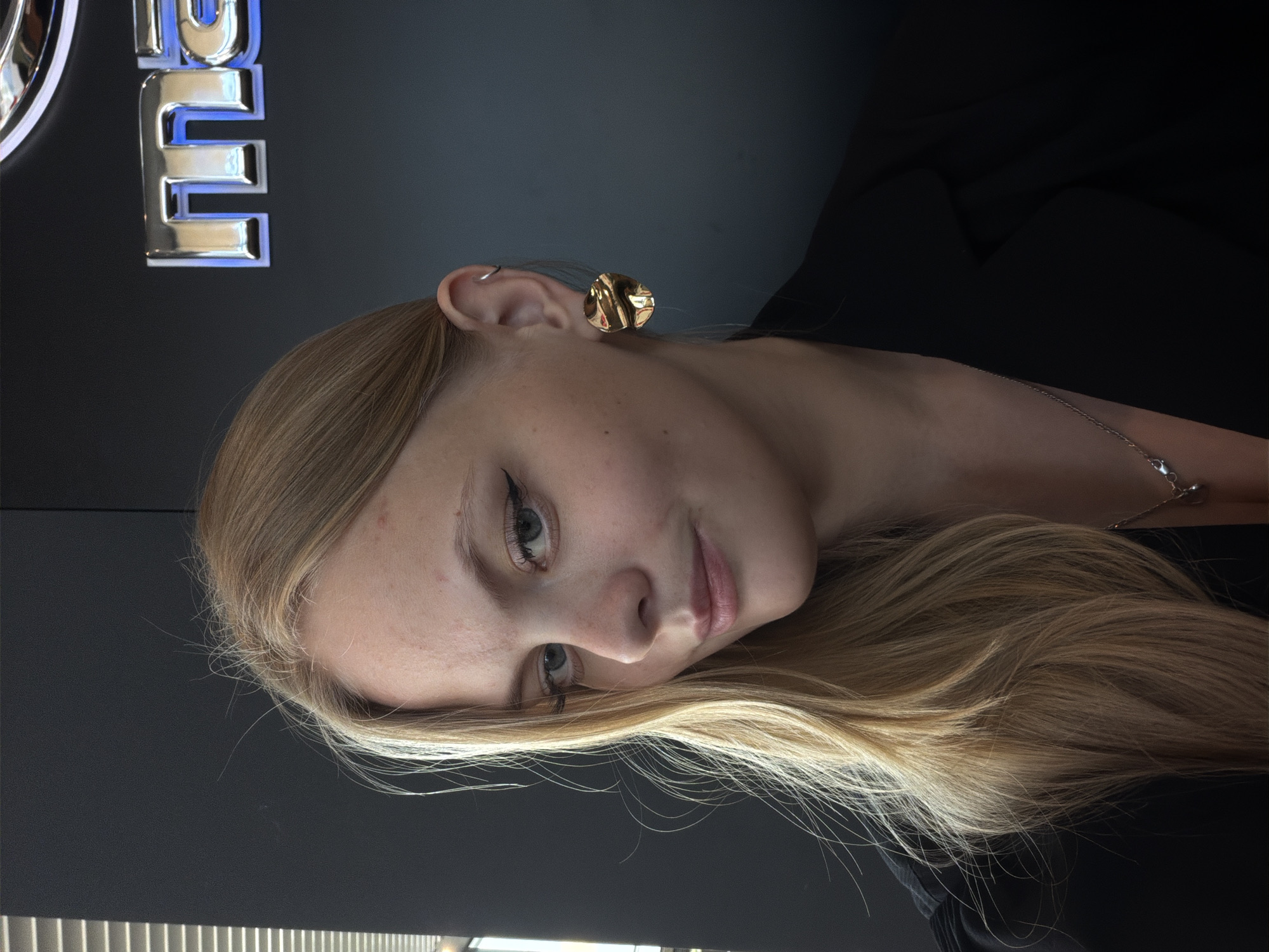 A fair-skinned woman with blond hair is positioned against a dark backdrop. She is wearing a gold earring in her right ear. A silver necklace is around her neck. Part of a sign is visible in the upper left corner of the image with a stylized "E" in blue and silver. The location cannot be determined from the image alone, and there is no way to place this photo in Izhevsk, Russia.