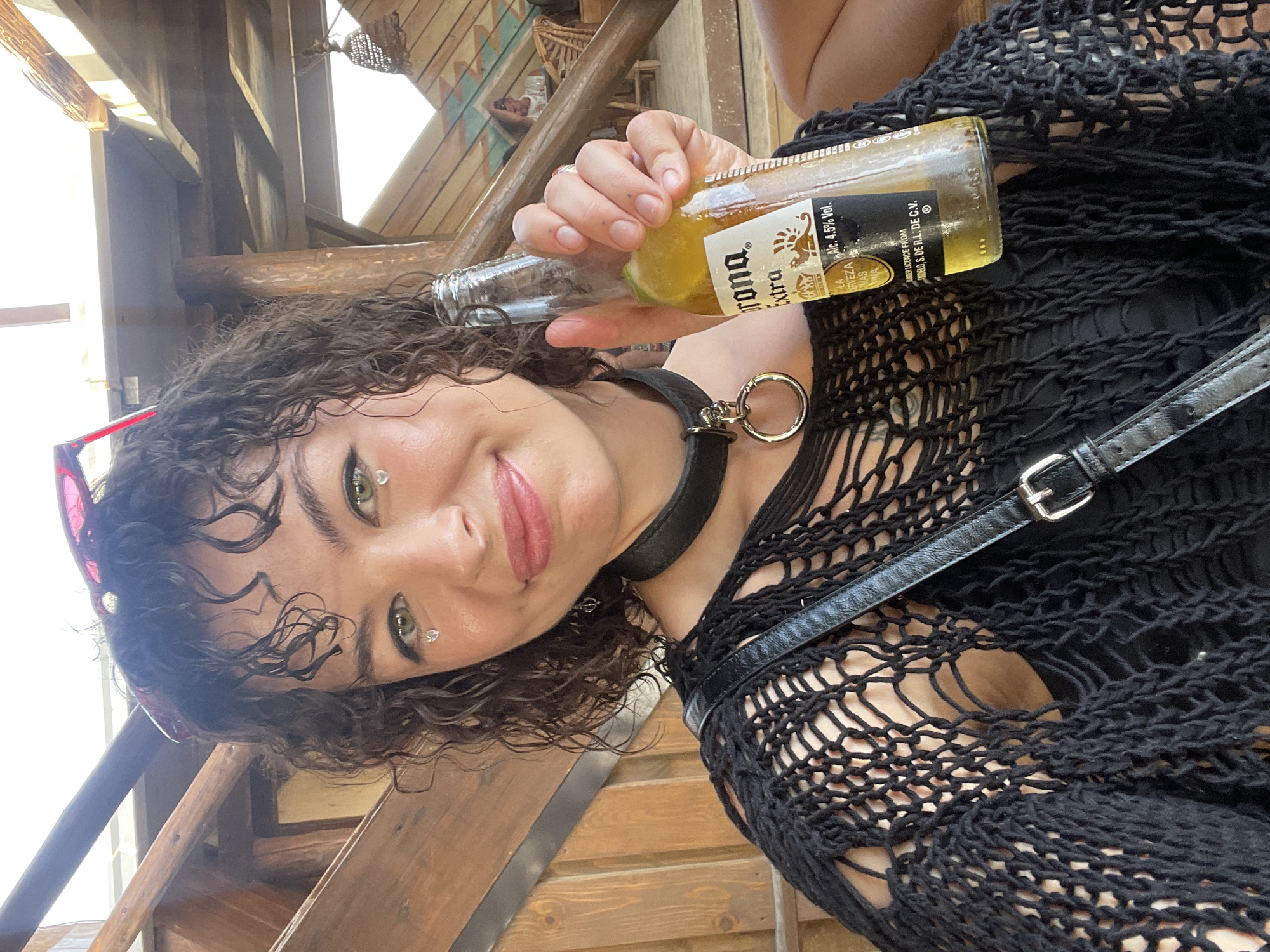 A young woman with curly dark hair and green eyes, adorned with heart-shaped glitter on her cheeks, smiles at the camera while holding a bottle of Corona Extra beer. She is wearing a black, mesh, off-the-shoulder top over a black tank top, and a black choker necklace with a silver ring. She is seated outdoors on a wooden structure, possibly a bar or restaurant, with wooden beams and railing visible in the background. In the distance, other patrons and seating areas can be seen. The lighting suggests it is daytime. The woman's pose and the beer suggest a casual, social setting. The location is identified as Donji Štoj, Montenegro.