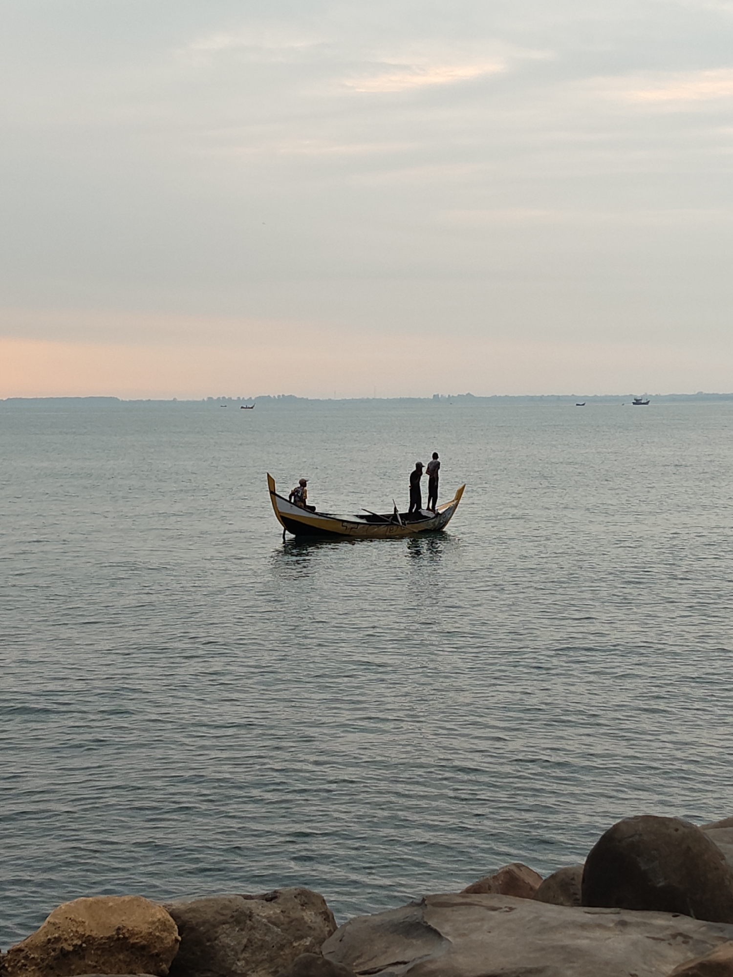 The image depicts a tranquil marine scene, likely at dawn or dusk, off the coast of Lhokseumawe, Indonesia. In the foreground, several large, weathered rocks form a partial barrier or shoreline. The midground features a calm body of water with subtle ripples. Centrally positioned on the water is a small wooden fishing boat, approximately 6-8 meters in length, with a black hull and prominent yellow trim along the gunwales, particularly at its pointed bow and stern. The number "187" is faintly visible on the bow. Three individuals are aboard the boat. One person is seated towards the bow, facing inwards, wearing light-colored clothing and possibly a head covering, appearing engaged with an object in the boat. Two other individuals stand upright near the stern, their figures silhouetted against the ambient light, both appearing focused on activities within the boat. In the distant background, a low-lying landmass or coastline is visible along the horizon. Several other much smaller vessels appear as dark specks on the distant water, with one larger, silhouetted vessel positioned further out to the right. The sky overhead is overcast or hazy, displaying a gradient from light peach and orange hues near the horizon to a pale grey-blue directly above.
