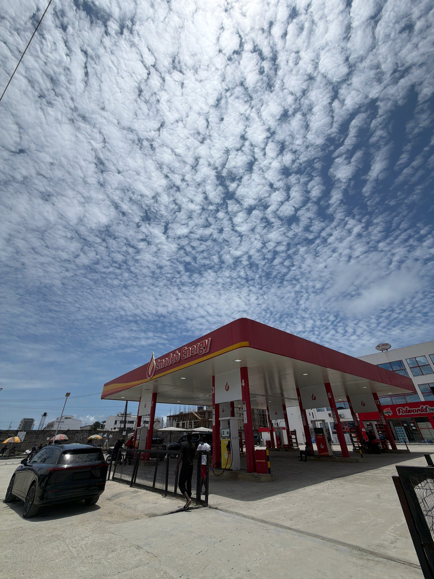 A street-level, wide-angle shot captures a brightly lit gas station under a vast, cloudy sky. The gas station, identified by the "Emadeb Energy" signage and red and yellow color scheme, features multiple fuel pumps under a covered canopy. A black SUV is parked to the left of the frame, angled slightly towards the viewer. Several individuals are visible around the gas station, some standing near the pumps and others walking. In the background, buildings, including one with "Polo World" signage, and a few scattered umbrellas suggest an urban environment. The ground is paved, with some shadows indicating a sunny day despite the clouds. The scene appears to be the Emadeb Energy gas station located in Igboefon, Nigeria.