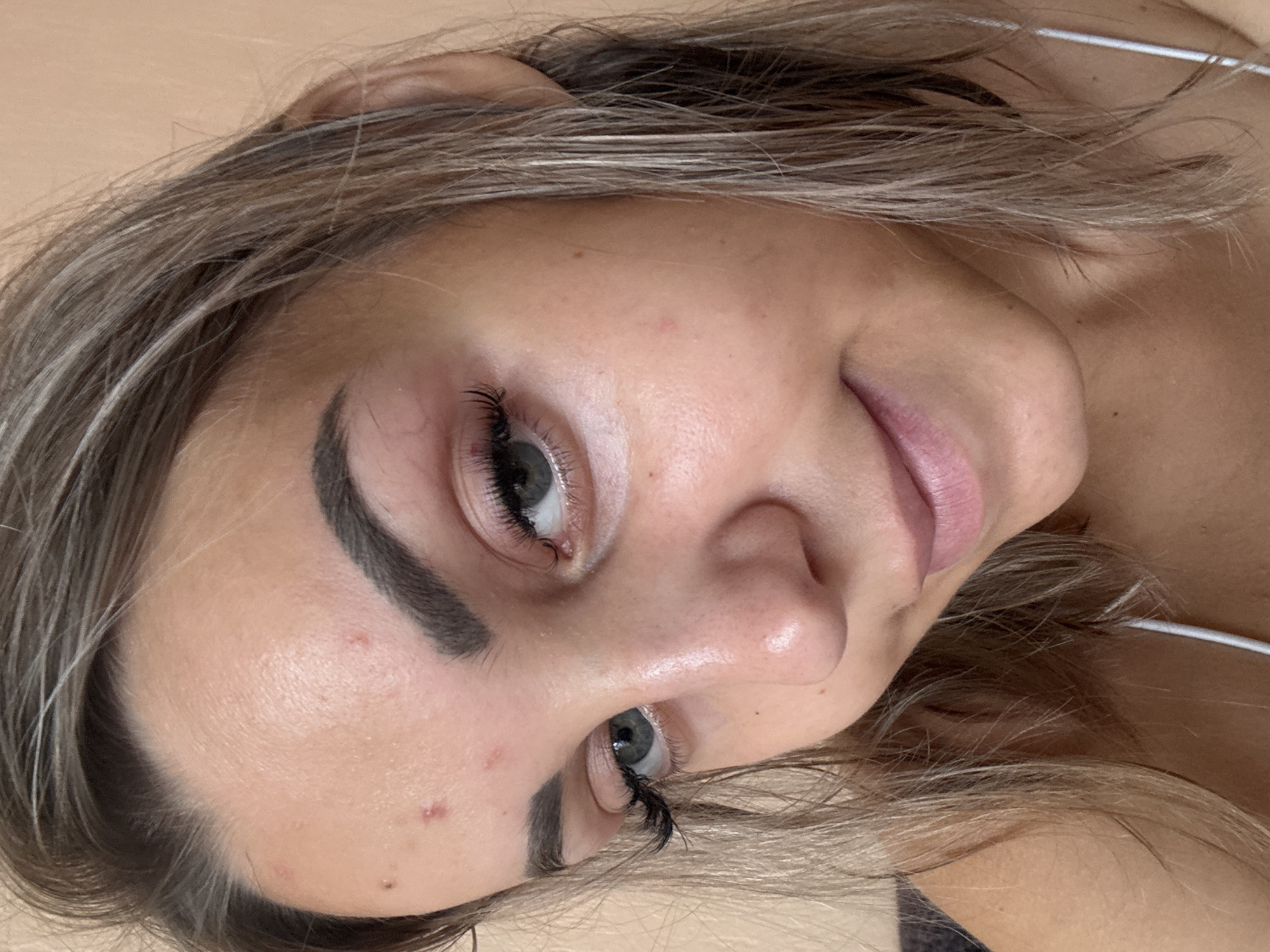 A close-up, waist-up, selfie-style photograph features a young woman with light brown hair, light eyes, and fair skin. Her hair is styled down and appears slightly damp. She is wearing a white strap top. The woman has noticeable blemishes, including several red pimples on her forehead and cheek. Her eyebrows are dark and well-defined, and she is wearing false eyelashes. The background is a plain, light peach or beige color, suggesting an indoor environment. The lighting is soft and natural, illuminating her face from the front. The photograph does not contain any elements that would allow for the determination of its location in Kholmsk, Russia, or any other specific city.