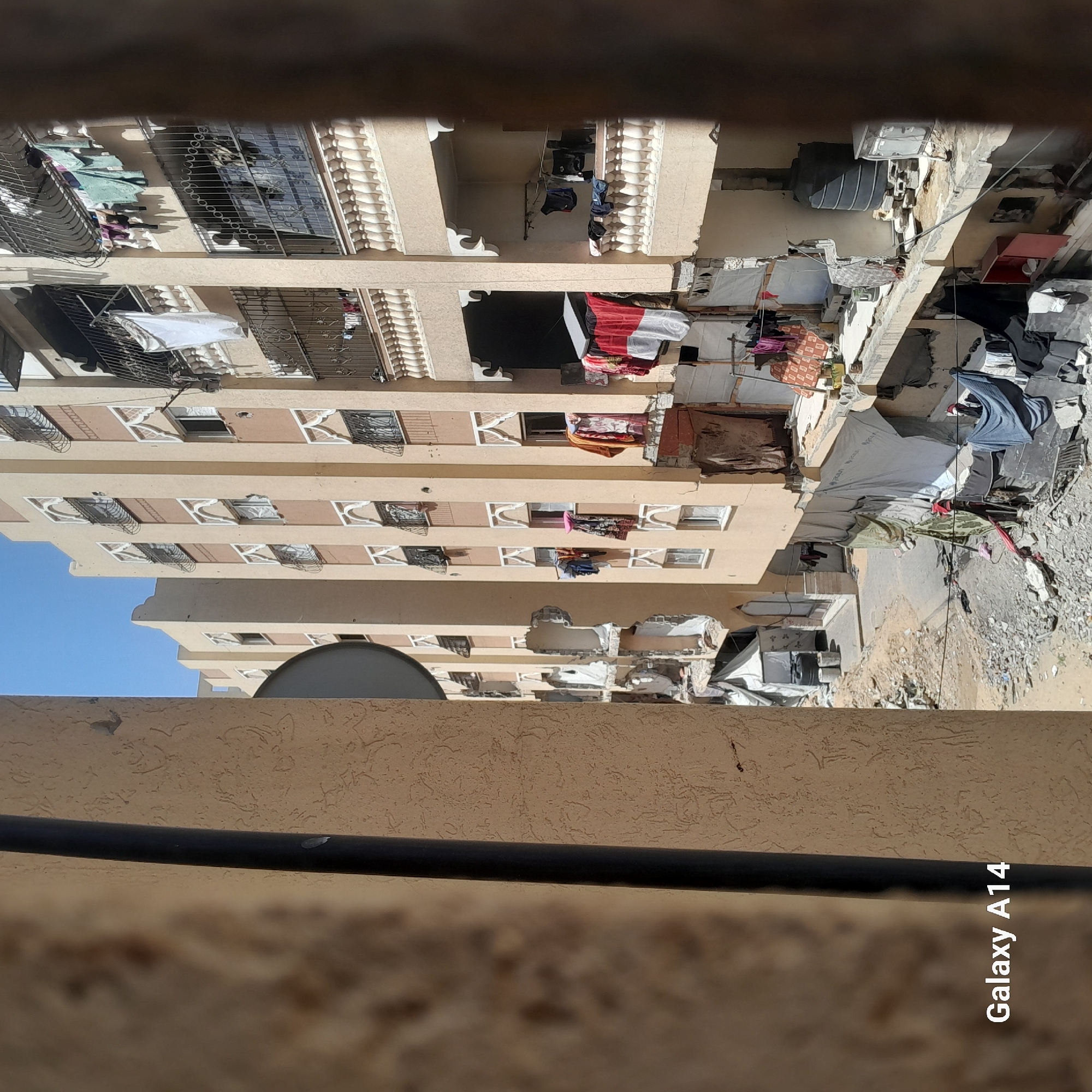 The image, taken from a high vantage point, depicts a multi-story building in a state of partial ruin. The building is primarily tan in color with repetitive window designs, some featuring decorative ironwork. Clothing is hanging from multiple balconies and windows. A satellite dish is visible on one of the lower levels. To the right of the building, there is a large pile of debris, suggesting a significant collapse or explosion. The ground is covered in rubble and dirt. A black wire or cable is visible in the foreground, partially obscuring the scene. The sky is blue. The location is Unknown, Unknown.