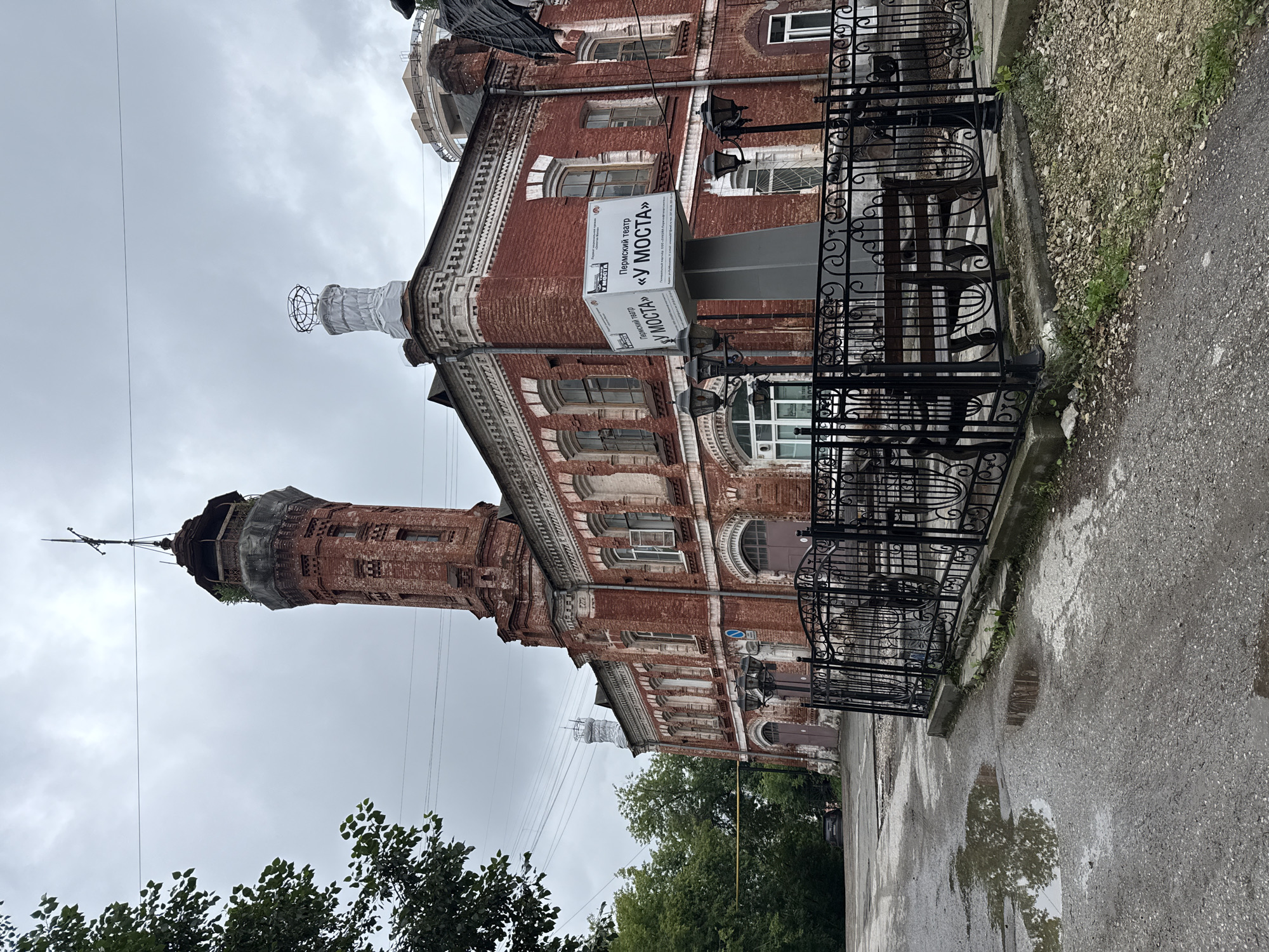 An overcast day in the city of Perm, Russia, features a prominent red brick building with a tall, weathered tower. The building has multiple windows and arched doorways, with decorative brickwork along the roofline. A modern sign stands in front of the building, displaying Cyrillic text and the name "У МОСТА" (U Mosta), which translates to "At the Bridge." The sign appears to be advertising the Perm Theater.
The area in front of the building is a paved courtyard, with several puddles reflecting the cloudy sky. A black wrought-iron fence encloses a small seating area with a wooden bench. Several outdoor lamps are visible along the fence. To the left of the building, a cluster of green trees partially obscures the structure. Power lines are strung across the sky above the building and trees. In the distance, a dark-colored car is parked on the street.