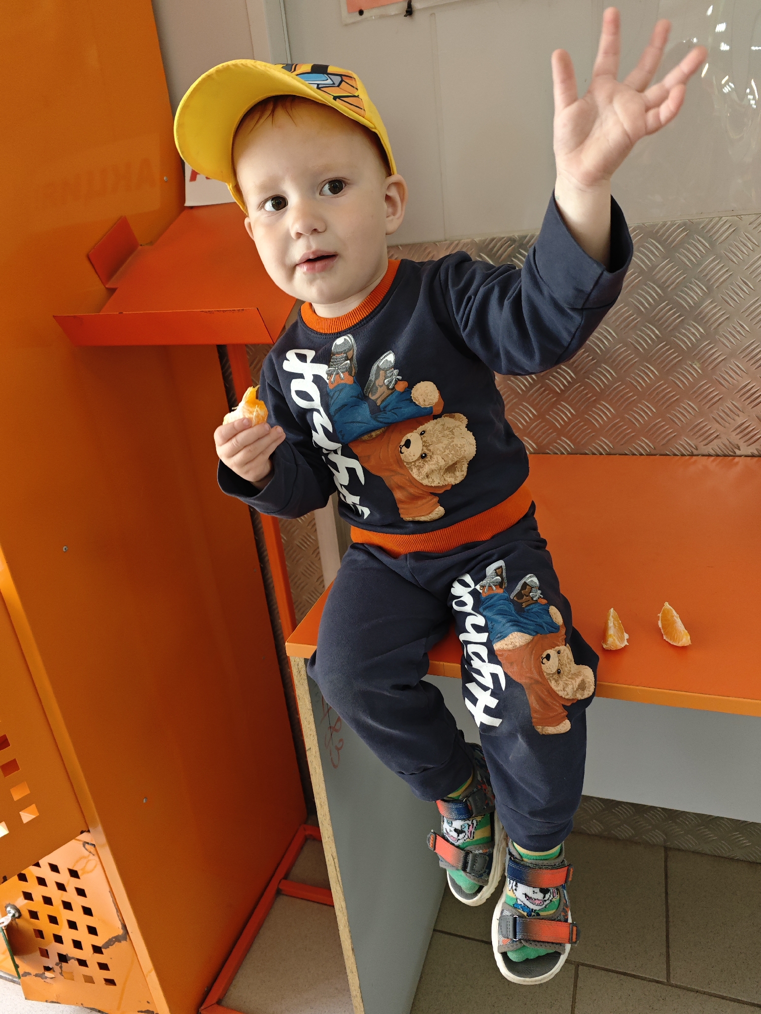 A young boy sits on an orange bench in what appears to be a public space. He is wearing a yellow cap, a navy blue sweatsuit with a bear graphic, and sandals with colorful socks. He holds a piece of orange in his left hand and raises his right hand, palm outward. Three orange segments are on the bench next to him. The location is the city of Chaikovsky, Russia. A section of metal flooring with a diamond pattern is visible behind the bench. Part of an orange structure with cut-out squares is to the boy's left.