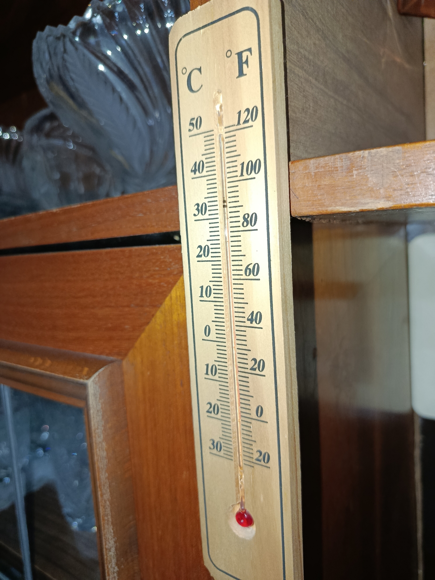An old wooden thermometer hangs on a wooden surface. The thermometer has markings for both Celsius and Fahrenheit. The Celsius scale ranges from -10 to 50 degrees, and the Fahrenheit scale ranges from 0 to 120 degrees. A red liquid is visible within the thermometer's glass tube, indicating the current temperature. In the background, out of focus, there are some shiny, silvery objects that appear to be decorative. The overall setting suggests an indoor environment, possibly a room or a cabinet.