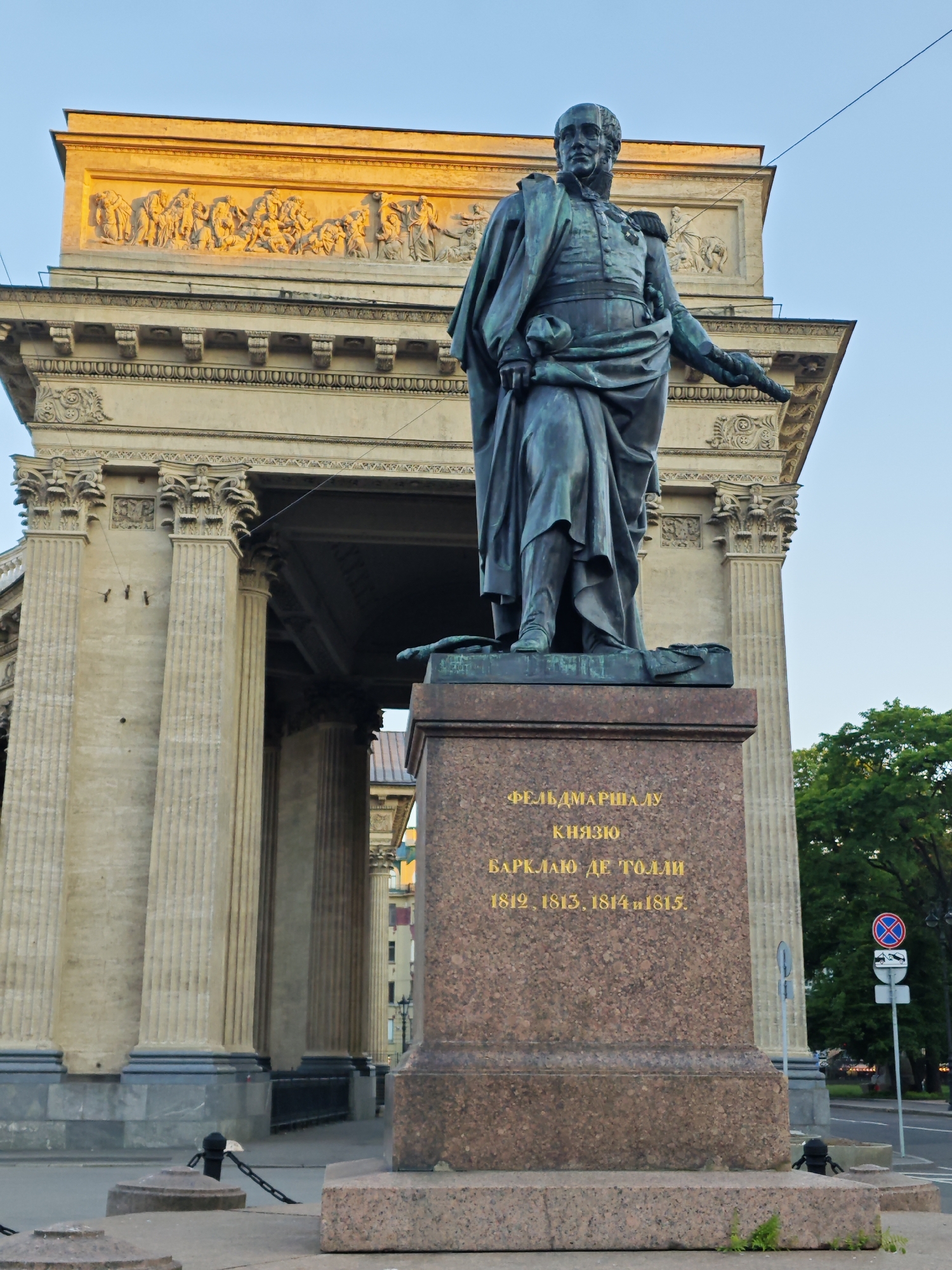 The image features the bronze statue of Field Marshal Prince Mikhail Barclay de Tolly positioned in front of the Kazan Cathedral in Saint Petersburg, Russia. The statue depicts a male figure with a greenish-blue patina, clad in military attire including a uniform with visible medals and epaulets, and a flowing cape. His left arm is bent while his right arm extends forward and slightly downward. The statue stands on a tall, rectangular pedestal made of reddish-brown granite with visible flecks. Inscribed on the pedestal in gold Cyrillic script is "ФЕЛЬДМАРШАЛУ КНЯЗЮ БАРКЛАЮ ДЕ ТОЛЛИ 1812, 1813, 1814 и 1815.". The pedestal is supported by a multi-tiered base of the same granite, with parts of a dark chain barrier and light-colored bollards visible in the immediate foreground.
Behind the statue, the neoclassical facade of the Kazan Cathedral dominates the background. The building, constructed from light-colored stone, features a prominent portico composed of numerous tall, fluted columns with ornate Corinthian capitals. Above the columns, a pediment houses a detailed bas-relief sculpture depicting multiple human figures in various poses. This relief is strongly illuminated by warm, golden sunlight originating from the right, creating a distinct contrast with the shadowed sections of the lower building. The sky above is clear and light blue. To the far right, green tree foliage is visible, alongside a segment of a street or pathway, a dark lamppost, and a traffic sign displaying a red circle with a diagonal line over a small car with a tow hook, indicating no stopping and potential towing.