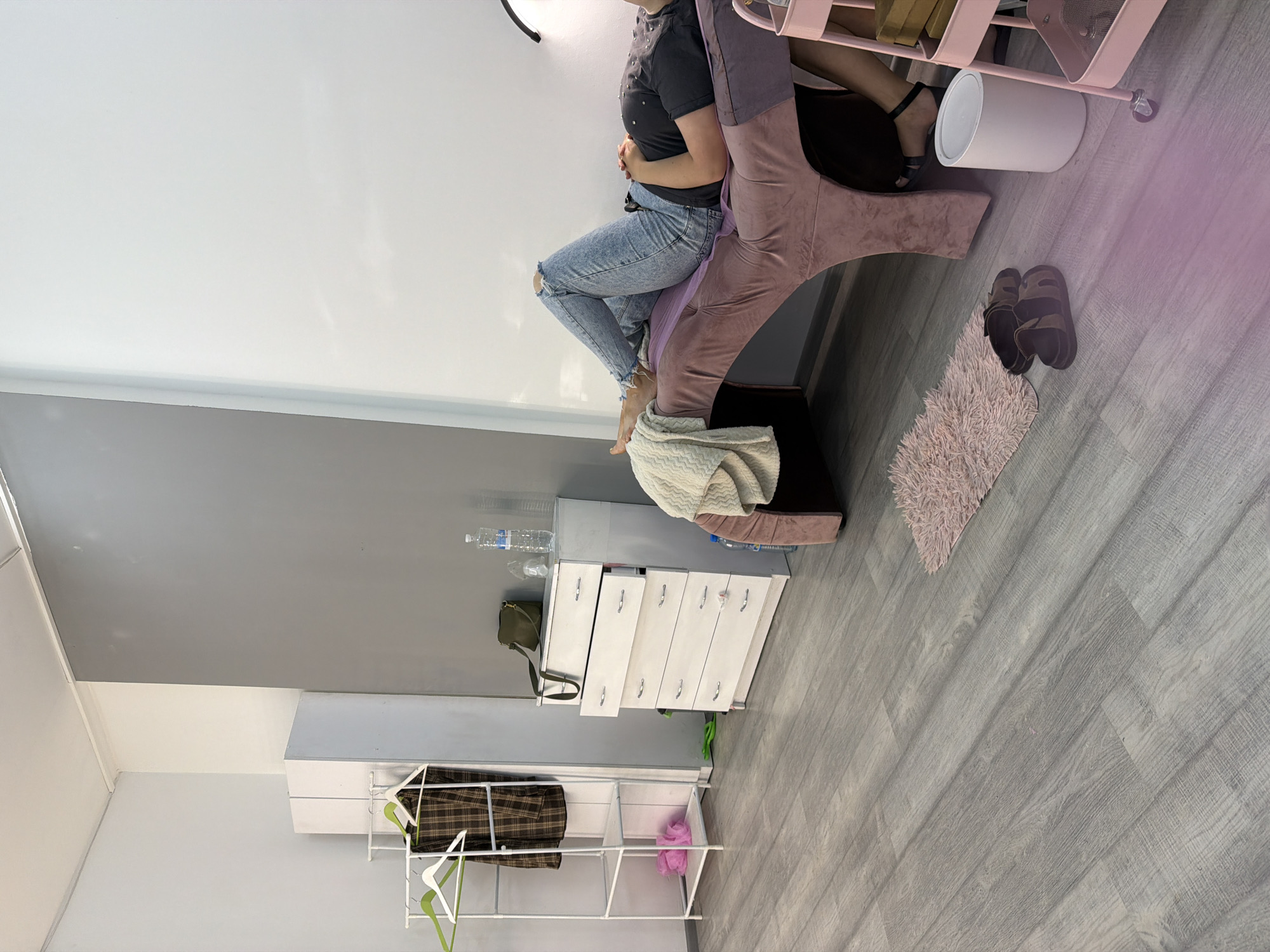 A person lies on a pink massage table in a room with gray and white walls. They are wearing ripped blue jeans and a dark t-shirt. To the left of the table, there is a white dresser with several drawers. On top of the dresser, there are two bottles of water and a green purse. Next to the dresser is a white clothing rack with a plaid shirt hanging on it. In the foreground, there is a small pink rug and a pair of brown sandals. A white trash can and a pink rolling cart with shelves are visible to the right of the massage table. The floor is made of gray wood-look laminate. The scene appears to be an indoor salon or treatment room.