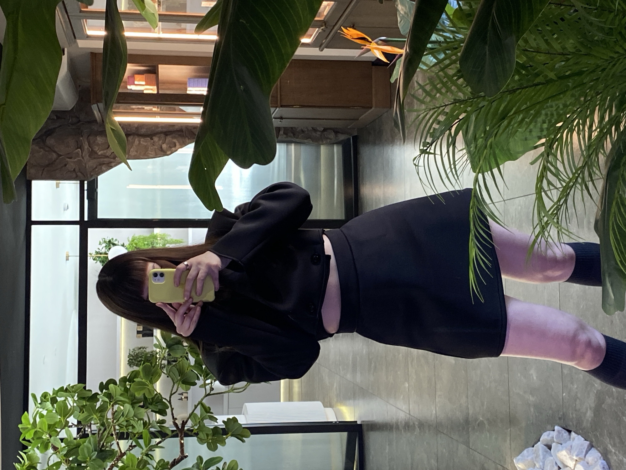 A person with long, dark hair is taking a selfie in a mirror. They are wearing a black skirt, black jacket, and black socks. The phone in their hand is yellow. The reflection shows a window with black frames, greenery, and a glimpse of a building exterior, a decorative wall texture, and interior lighting. The location is not explicitly identifiable as Votkinsk, Russia, from the provided image.