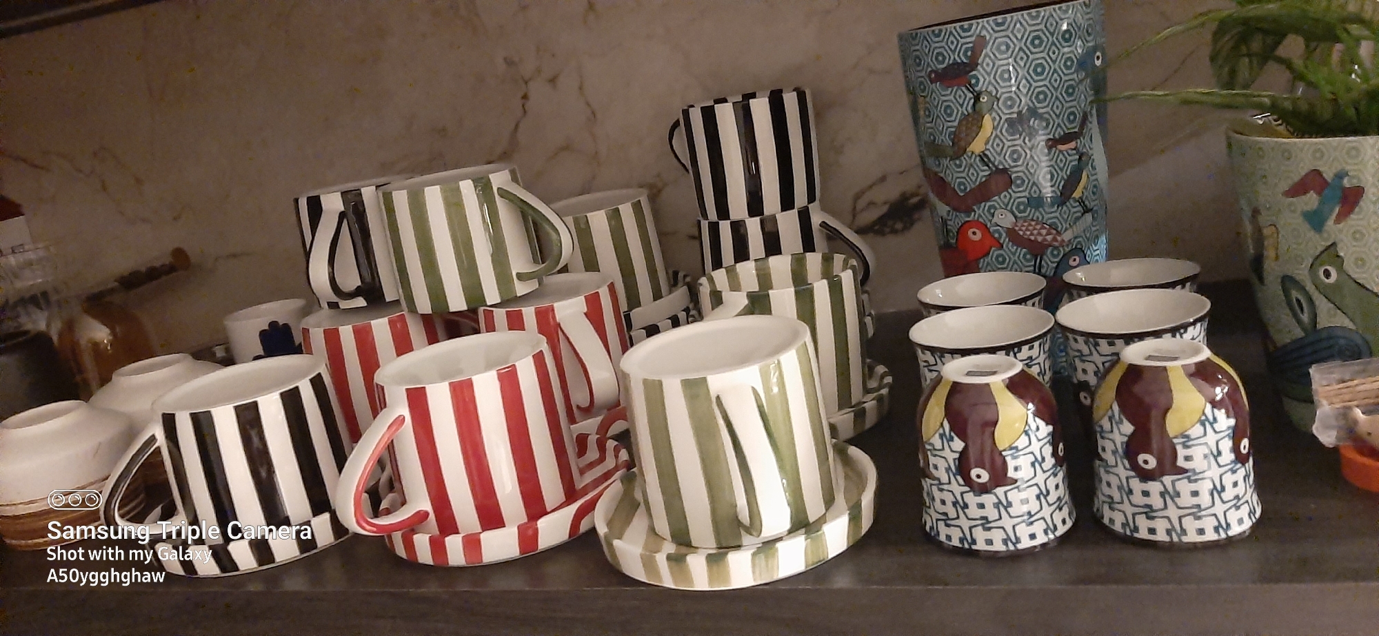 The image displays a collection of ceramic mugs and a vase, likely on a shelf or counter. The objects are primarily decorated with vertical stripes in various color combinations: black and white, red and white, and green and white. Some mugs are stacked, while others are arranged individually with their saucers. To the right, a large vase features a colorful pattern of birds and geometric shapes. In the background, a plant is visible, adding a touch of greenery. The overall scene appears to be indoors, possibly in a home or shop, with neutral-colored marble-patterned walls behind the displayed items. The lighting suggests it is daytime, but no specific time can be determined. The image also contains visible text in the bottom left corner: "Samsung Triple Camera Shot with my Galaxy A50yggghghaw". There are no people or animals visible in the image, aside from those depicted on the vase.