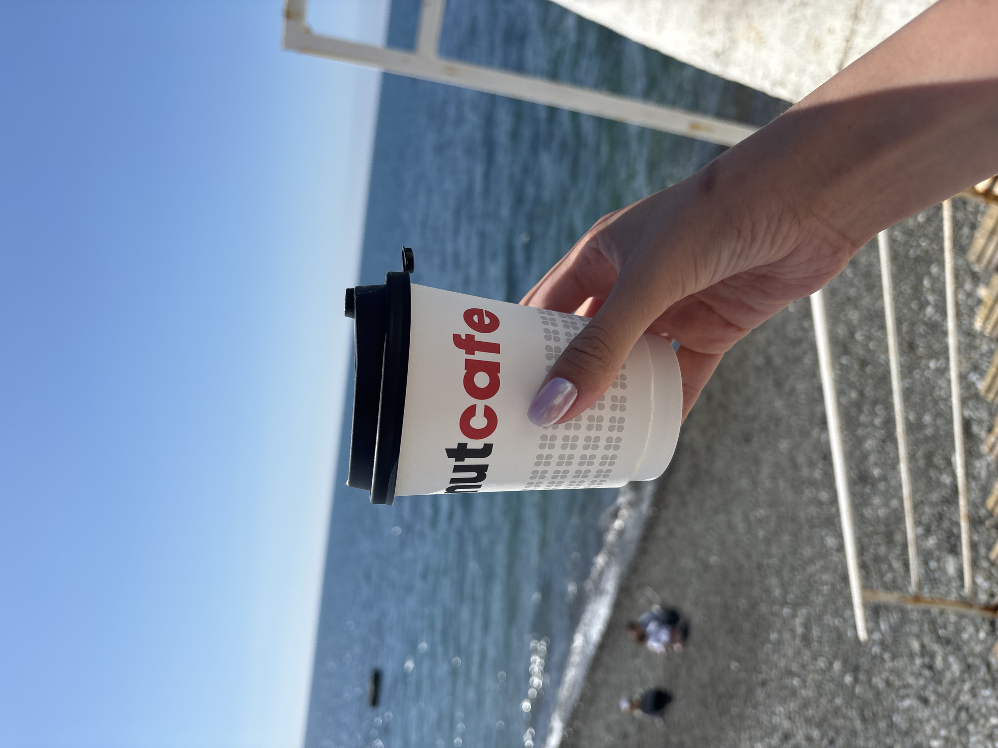 A hand holds a cup of coffee with the logo "nutcafe" against the backdrop of the Black Sea, likely in Sochi, Russia, suggested by the coastal environment and architecture present. The sky is clear and blue, suggesting a sunny day. The cup features a black lid and a pattern of gray icons beneath the brand name. People are visible in the distance on the beach. The edge of a man-made structure (likely a pier or railing) is partially visible in the foreground.