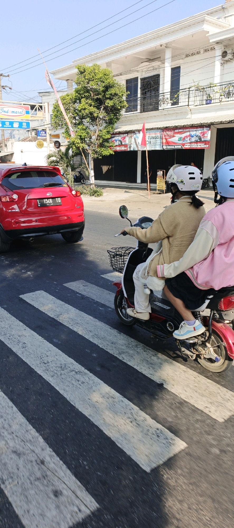 A red Nissan Juke, license plate L 1546 TD, is stopped at a pedestrian crossing on a paved road. Two individuals wearing helmets are riding a red and black electric scooter behind the car, also at the crossing. The person in front is wearing a light brown sweater and white pants, while the person in the back wears a pink and light blue jacket. They are both looking forward. In the background, a two-story white building with balconies and dark-framed windows is visible. A tree with green foliage stands to the left of the building. Signs for "Indomaret," "ATM BCA," and "Tunggal Motor" are present in the mid-ground. The sky is clear and blue, indicating it is daytime. The scene is in Gumukmas, Indonesia.