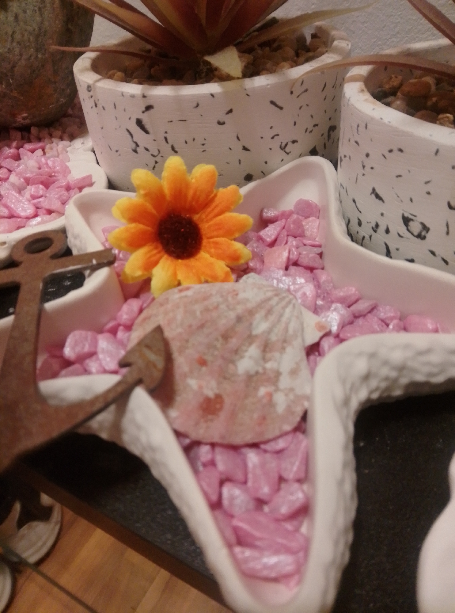 A decorative display features a white, speckled planter with a plant whose leaves are reddish-brown and extend upwards. To the right of this planter is another white, speckled planter containing small, brown stones and what appears to be the base of a plant. In the foreground, a white, star-shaped dish is filled with small, pink decorative stones. Nestled within these stones is a bright orange artificial flower with a dark center, and a light pink, oval-shaped seashell. To the left of the star-shaped dish, a rusty-brown anchor is positioned. The background shows a portion of a dark green textured pot or planter. The scene appears to be indoors, possibly on a shelf or table, with a wooden floor visible in the lower portion of the image. The overall impression is one of a decorative arrangement, likely a home or shop display.