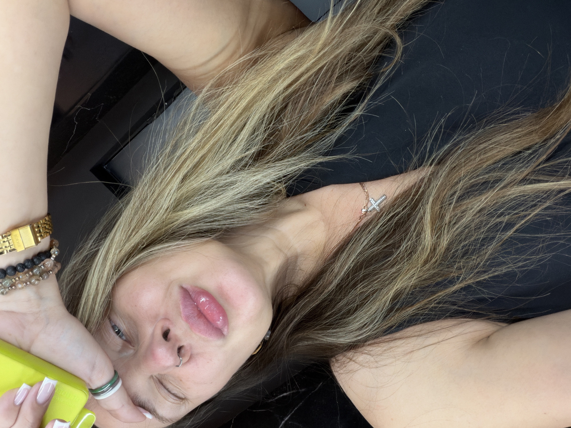 The image shows a close-up, selfie-style shot of a woman lying down and looking towards the camera. She has her eyes partially closed and her lips are pursed, with lip gloss on. She is wearing a black top, a cross necklace, a nose ring, and multiple bracelets on her wrist, including one with beads and a gold watch. She is also wearing several rings on her fingers, one of which appears to be a stack of green and white bands. Her hair is long and appears to be blonde with lighter streaks. She is holding a bright yellow object, possibly a phone case, near her forehead. The background is dark and out of focus, with some reflective surfaces visible. There is no information about the location within Укромновское сельское поселение, Ukraine, or any specific details about the environment.