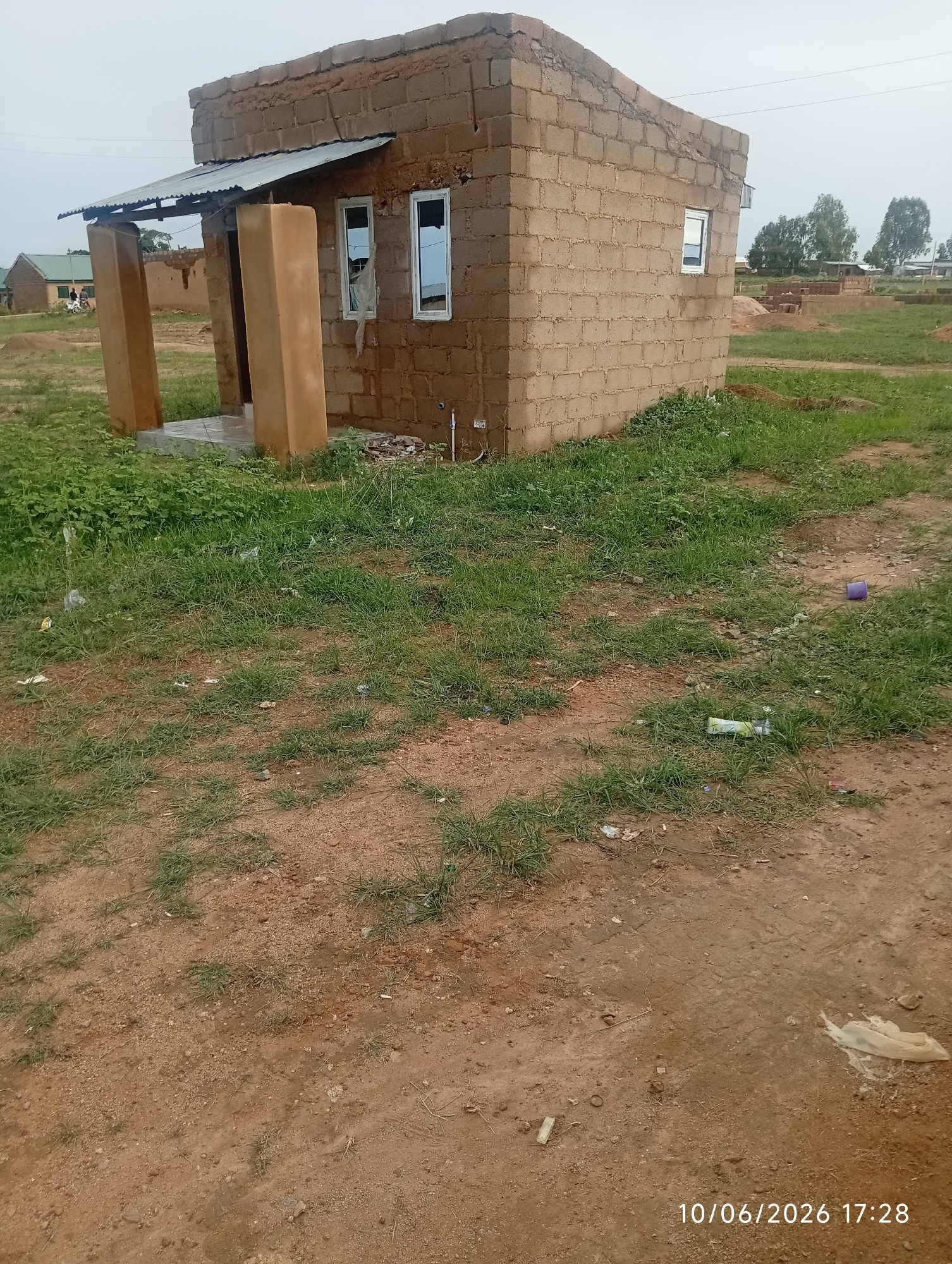A medium shot shows a partially constructed building with tan cinder blocks, a corrugated metal roof extension over the entrance, and two tall rectangular concrete columns supporting the roof. The building has white-framed windows. The surrounding area is an open field with dry brown dirt and patches of green grass, some of which are littered with plastic bottles and wrappers. In the background, other unfinished structures and trees are visible. The image is stamped with "10/06/2026 17:28".