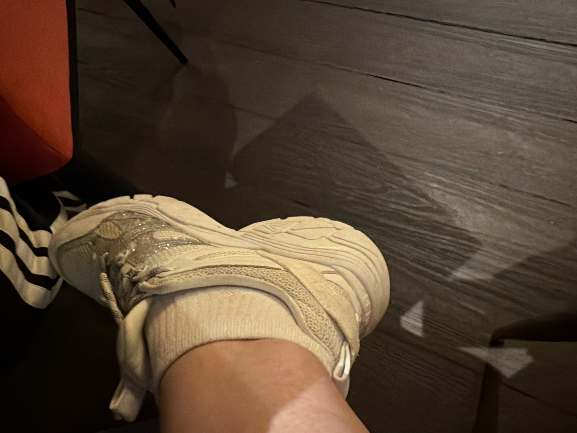 A person's foot clad in a beige sock and a white sneaker rests on a dark wooden floor. The scene appears to be indoors, and a partially visible red chair is located to the left. A small part of a garment with black and white stripes is visible next to the chair. The location is unknown, but Burgas, Bulgaria, has not been confirmed.