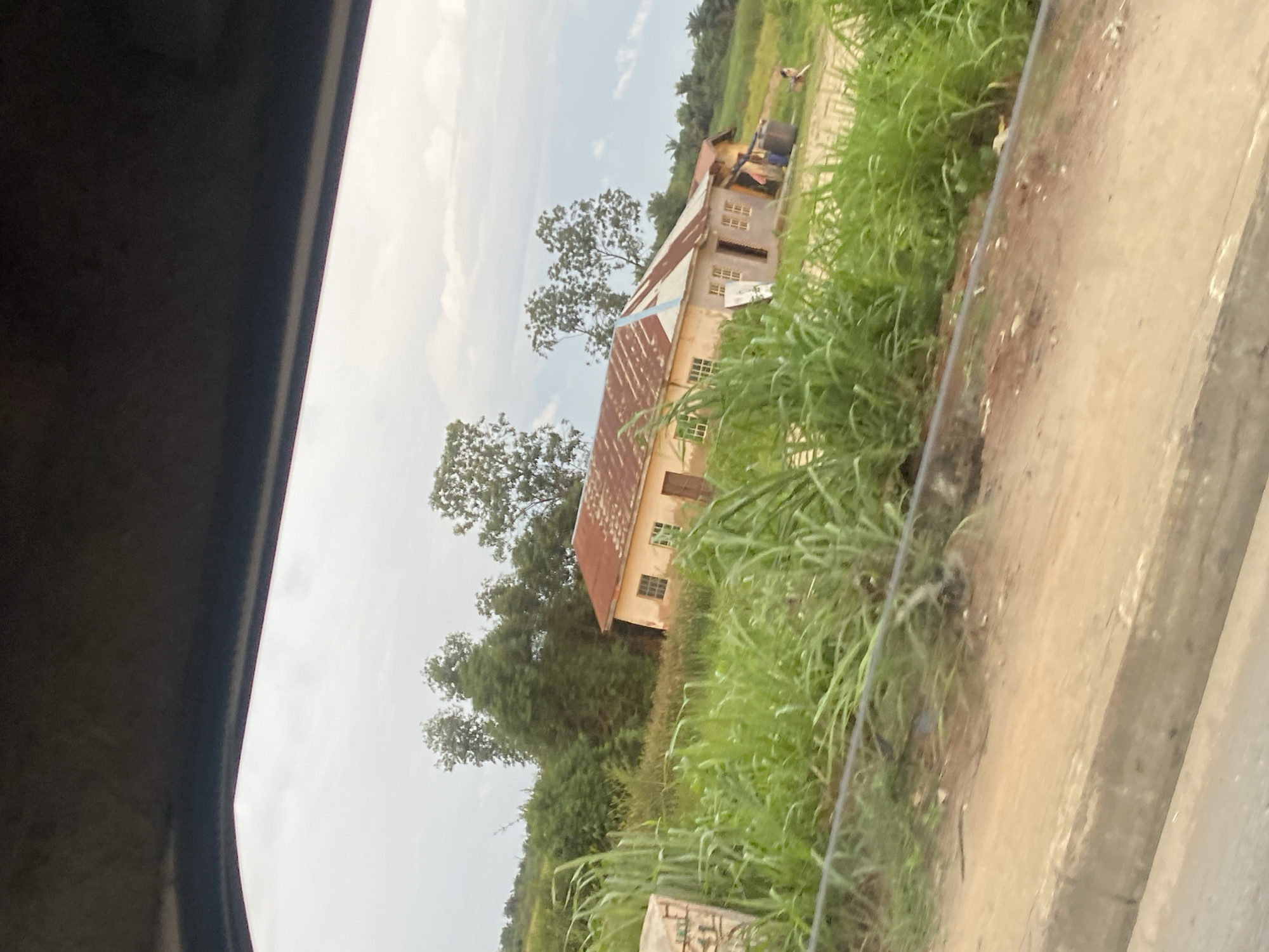 The image depicts a scene in Lagos, Nigeria, with a focus on a modest building and its surroundings. The building appears to be a single-story structure with a reddish-brown, corrugated metal roof. Its walls are a light color, possibly concrete, and there are several windows visible, some of which appear to have glass panes. The architecture suggests a functional rather than ornate design, possibly a school, community center, or a simple residence.
In front of the building, lush green grass and tall reeds grow densely, creating a natural border. A dirt path runs alongside the grass, leading into the distance. In the background, more trees and foliage are visible, hinting at a semi-rural or peri-urban setting within Lagos. The sky is overcast with clouds, suggesting a hazy or possibly rainy day, common in tropical climates. The lighting is diffused, indicating it could be morning or late afternoon.
There are no people clearly identifiable in the foreground or clearly engaged in any specific activity. However, in the distance, a figure can be faintly seen near some barrels, possibly indicating some form of work or activity associated with the building or its environment. The overall impression is one of a quiet, possibly slightly neglected, but functional space. The image is captured from a slightly elevated perspective, perhaps from inside a vehicle, with a dark, curved structure in the foreground suggesting a window frame or part of the vehicle's interior. No discernible text is present in the image.
