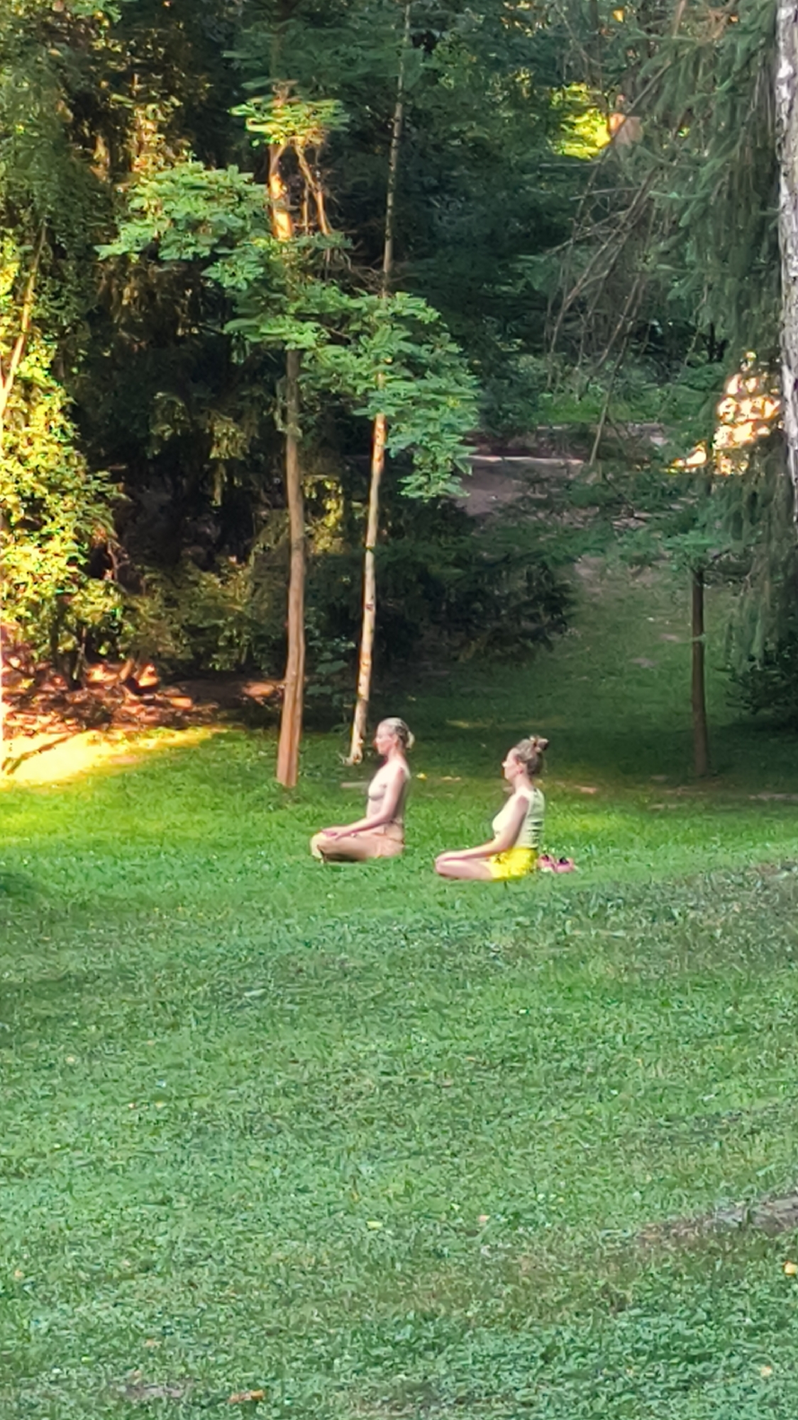 Two individuals are seated in a cross-legged meditation pose on a grassy lawn in a park. The person on the left is wearing a light-colored top and bottom, while the person on the right is wearing a light yellow top and yellow shorts. Both have their eyes closed. They are surrounded by lush green grass and trees, with sunlight filtering through the foliage, creating dappled light on the ground. The setting appears to be a peaceful, outdoor environment. The provided context states the image is from Lviv, Ukraine, suggesting this is a park within the city.