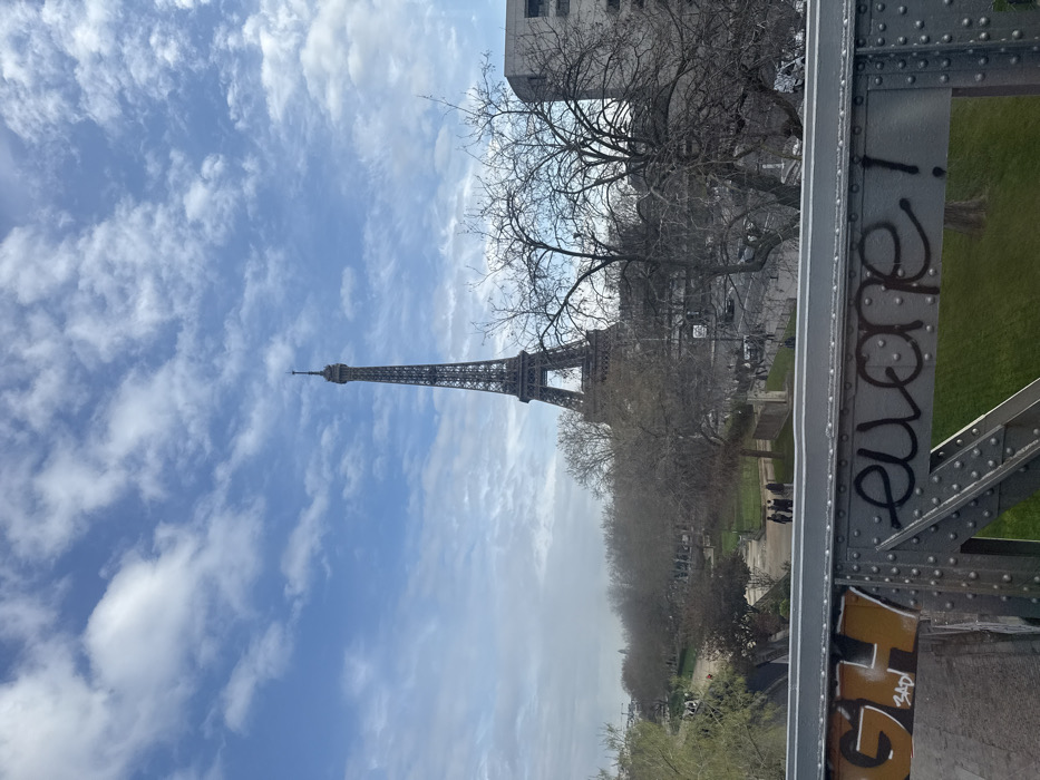 The image displays a panoramic view of the Eiffel Tower in Paris, France, observed from an elevated position on a metallic bridge structure. The foreground features a dark gray riveted metal bridge railing with graffiti; specific markings include "GH" in orange and "BAD" in black text on the left, and "euone !" in black text on the right. Below the bridge, a public park with green grass and numerous trees, predominantly bare of leaves, extends towards the left and center. Several small human figures are discernible walking on the park's pathways. To the right of the park, a multi-lane street is visible, with vehicles present, running alongside additional trees and modern-style buildings. The Eiffel Tower, a prominent iron lattice structure, dominates the background, rising against a bright blue sky dotted with scattered white and light gray clouds. The lighting suggests daytime conditions.