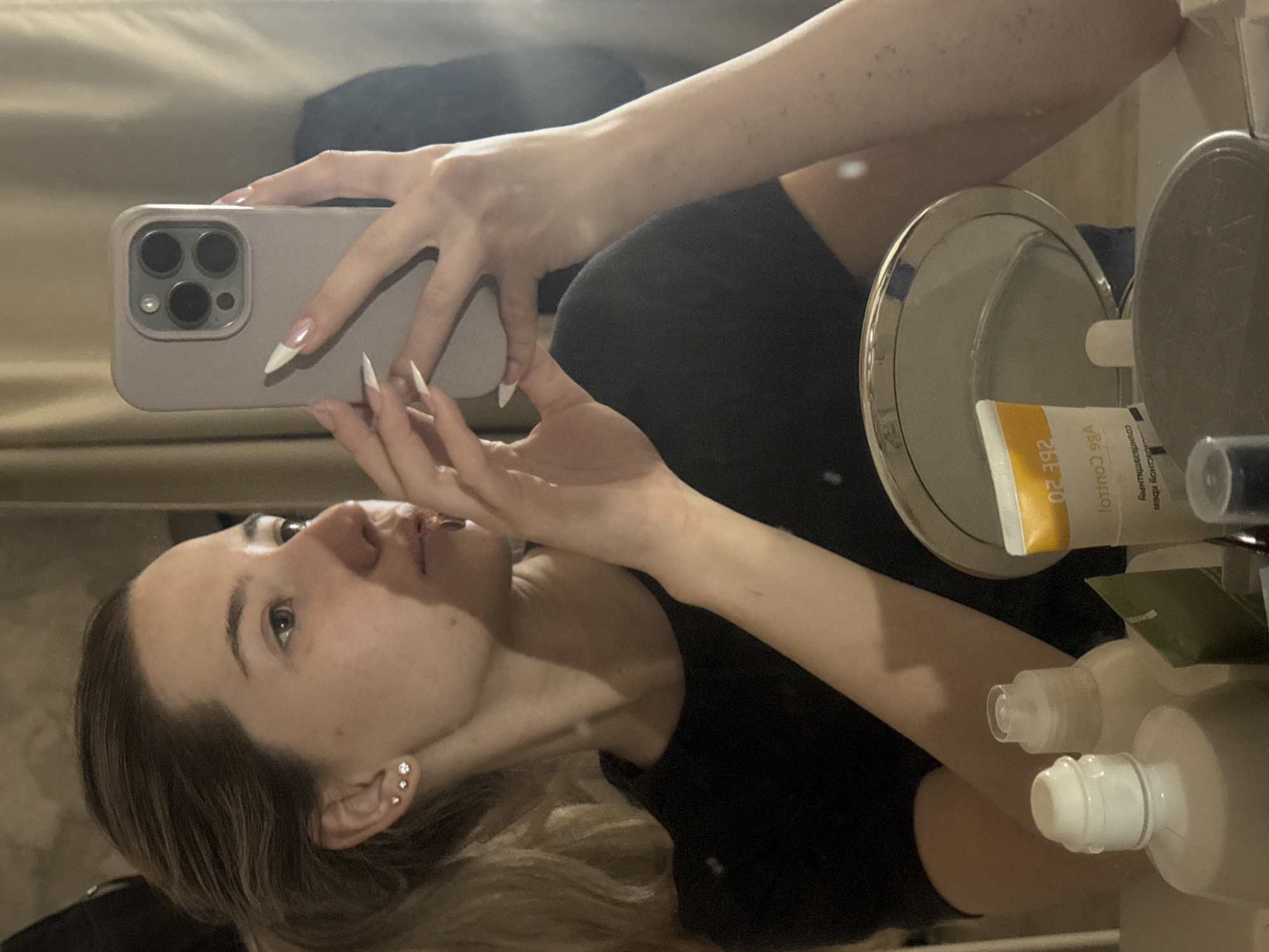 A young woman, likely in her late teens or early twenties, is captured in a mirror selfie taken indoors. She has light brown or blonde hair and is wearing a dark top. Her left ear features three small stud earrings. She is holding a smartphone in a light pink/purple case, which has a distinctive triple camera lens setup on its back. Her hands display long, pointed nails with a white French manicure style.
The woman is looking slightly upwards, with a neutral expression, presumably focused on her reflection or the phone screen while taking the picture. The setting appears to be a bathroom or bedroom, featuring a vanity area with a mirror. On the surface, a smaller round mirror with a metallic rim is visible, alongside several cosmetic or toiletry products. One yellow and white tube shows reversed text, including "0S 29E" (likely "SPF 20") and "loihtno3 qzA" ("Az Control"). Two opaque white/beige bottles with dispenser tops are also present. The background behind the woman is a light, neutral-toned wall or curtain. The lighting suggests an indoor setting, potentially evening or night. The image was taken in Noviy, Russia.