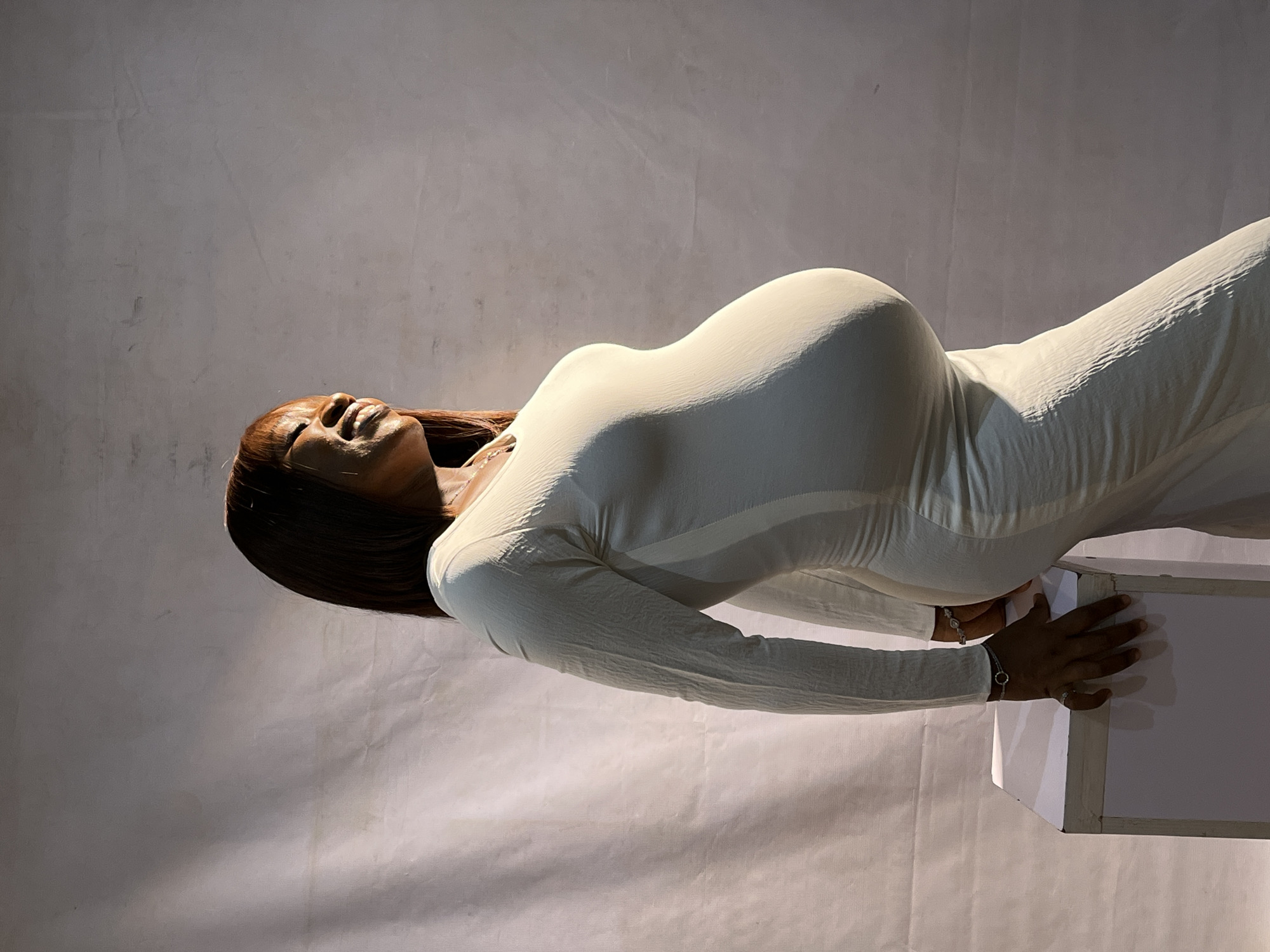 A pregnant woman, positioned in profile, stands in a studio setting. She is wearing a long-sleeved, off-white dress that accentuates her pregnant belly. Her hair is dark and cut into a bob. The woman's head is tilted back slightly, and she appears to be smiling with her eyes closed, bathed in dramatic lighting that casts shadows on the white backdrop. To her left, a white cube-shaped prop is visible, with her left hand resting on its surface. She is also wearing bracelets on her left wrist and a ring on her left index finger. The studio environment is minimalist, with a textured white backdrop. This image was taken in Jos, Nigeria.