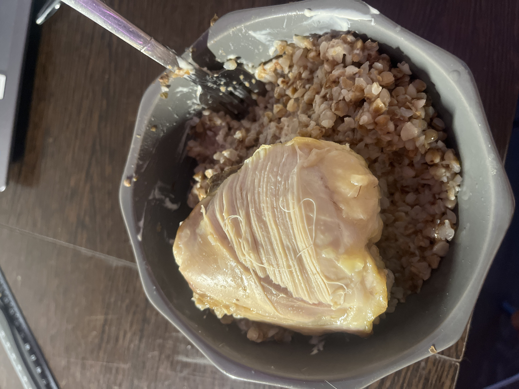 A close-up, overhead shot depicts a gray bowl filled with buckwheat and a piece of cooked chicken. The chicken sits atop the buckwheat, dominating the center of the bowl. A utensil, possibly a fork or spoon, is partially visible in the upper left corner of the frame. The bowl is on a dark, wooden surface. The lighting is somewhat uneven, creating shadows and highlights across the scene. There is no evidence of Saint Petersburg, Russia.