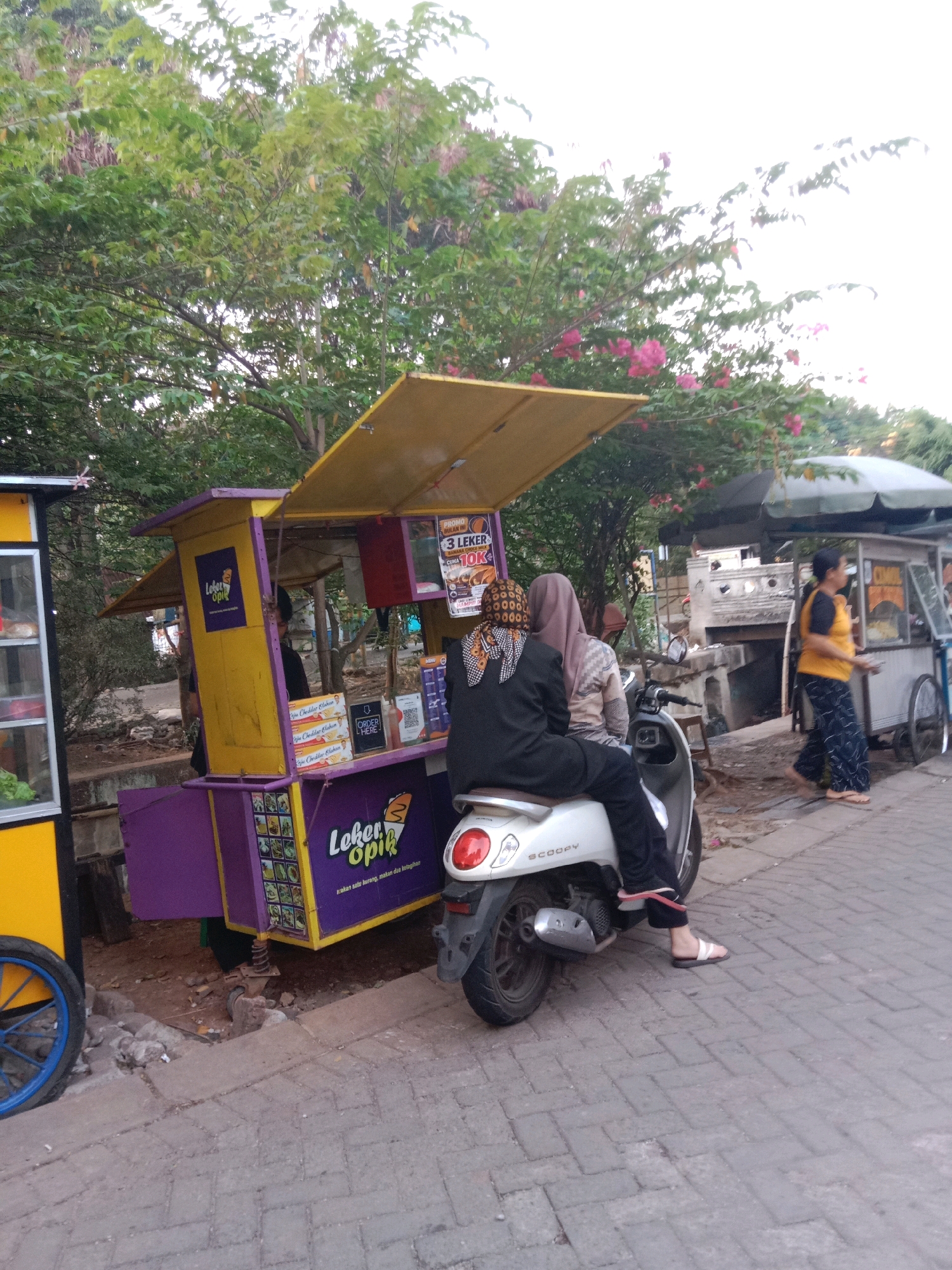 The image depicts a street scene in South Tangerang, Indonesia, with a focus on a food stall and its customers. A purple and yellow food cart, labeled "Loker Opik," is prominently displayed, suggesting it sells snacks or drinks. Two women, one wearing a black hijab and the other a lighter colored hijab, are seated on a white scooter parked in front of the food stall. The woman in black is wearing a black long-sleeved shirt and pants, and white flip-flops. The woman behind her is wearing a patterned long-sleeved shirt and a light purple hijab.
To the left of the food stall, another yellow cart with a glass display is partially visible, possibly another food vendor. In the background, a woman in an orange top and dark patterned pants is walking away from a different food cart. This cart is larger and appears to have a green canopy. The environment is characterized by lush green trees with some pink blossoms, and the ground is paved with bricks. The overall impression is a busy street with mobile food vendors operating in an outdoor setting.
